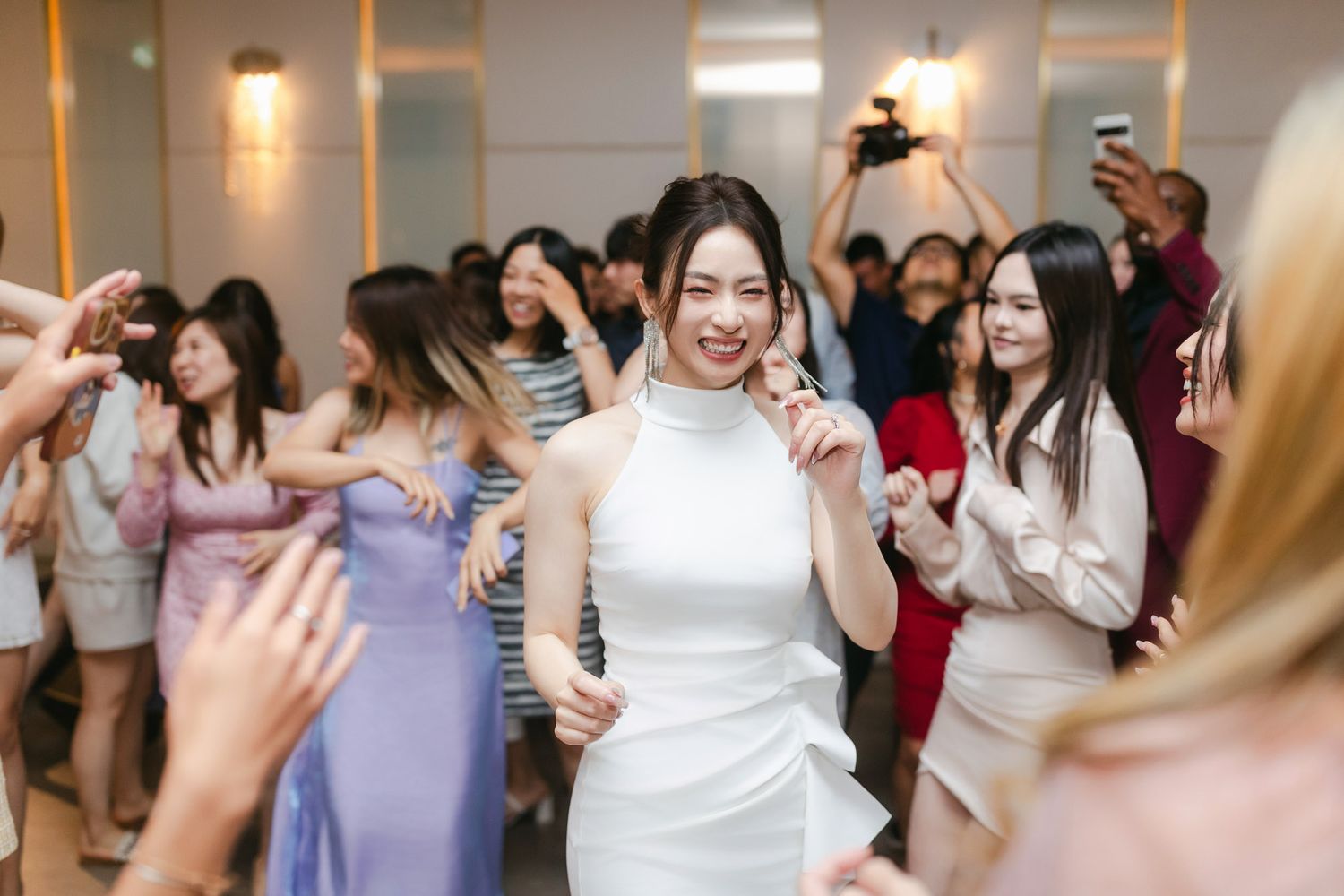 Bride in white halter gown dancing joyfully surrounded by guests at a wedding reception celebration.