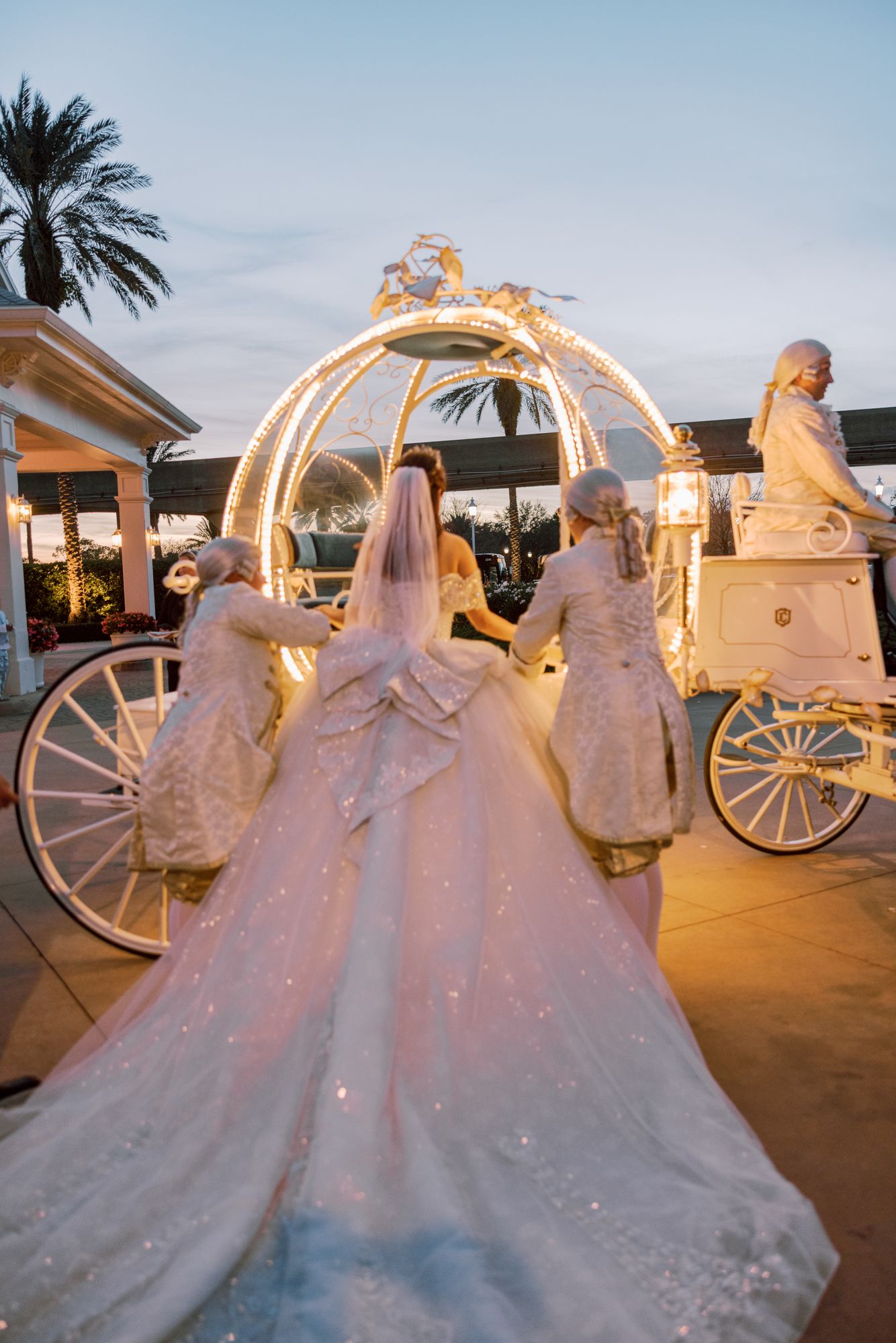 Bride in sparkling white gown approaches a glowing Cinderella carriage with attendants at dusk.