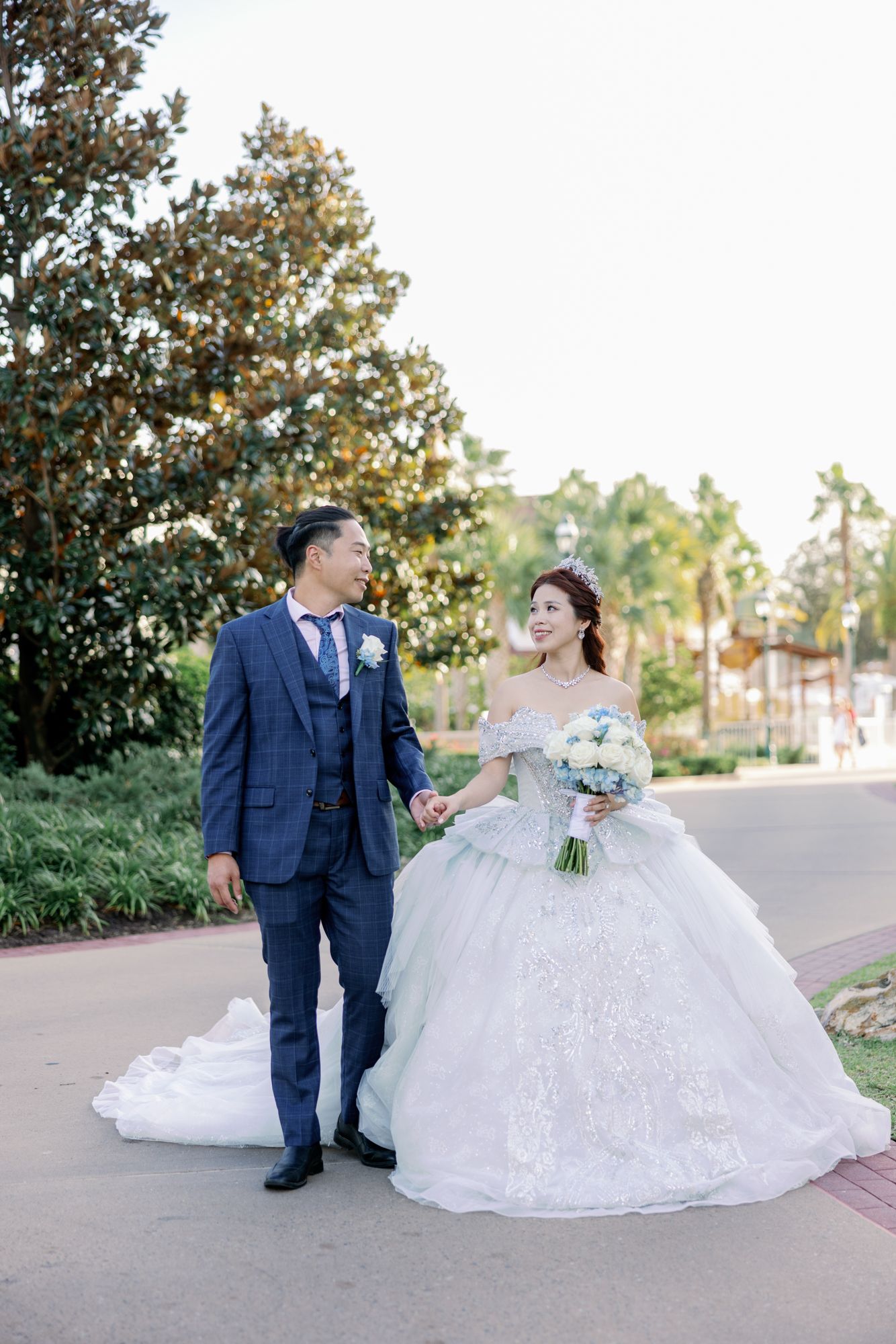 Asian bride and groom holding hands in garden, bride in off-shoulder ball gown with bouquet.