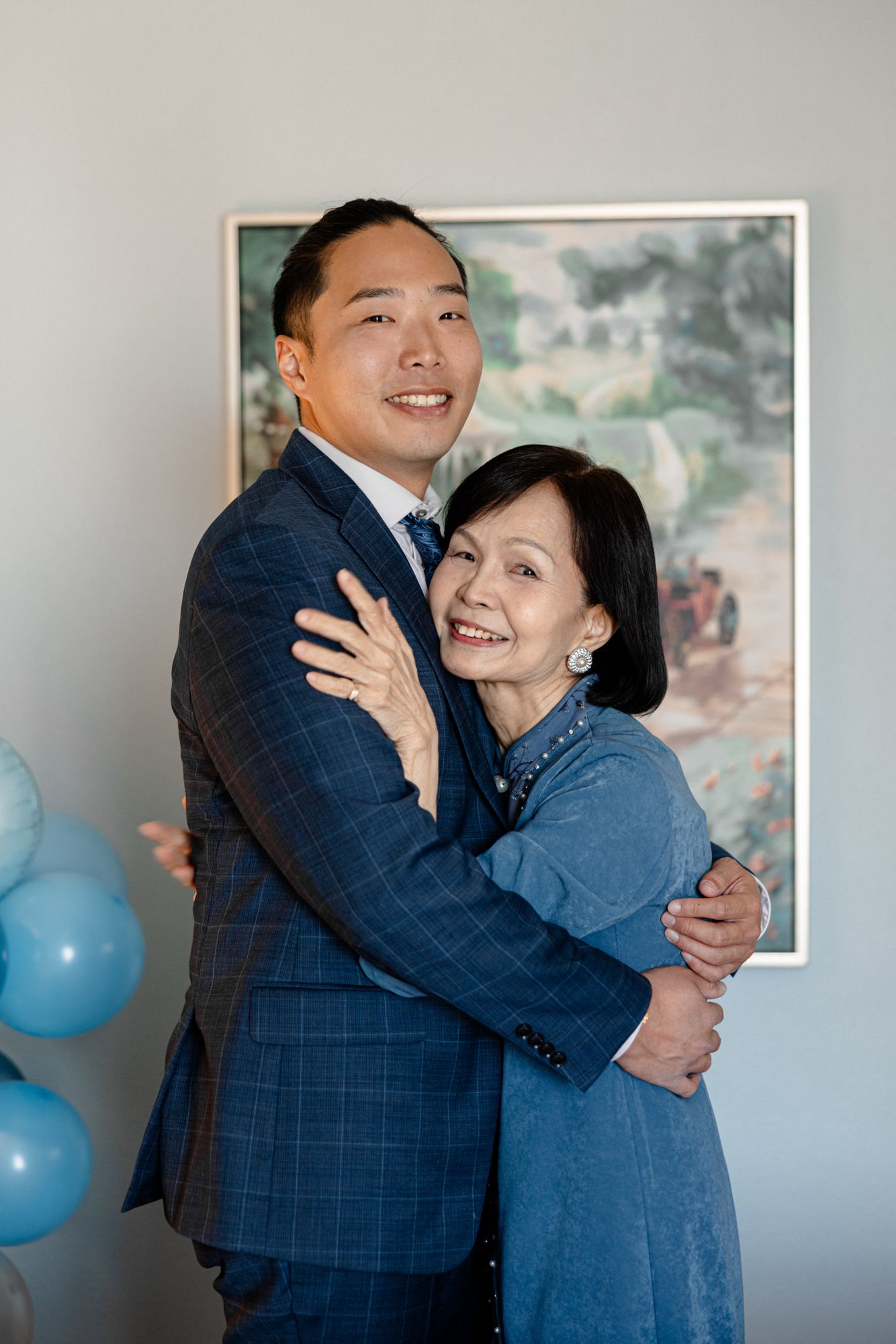 A smiling Asian couple in blue attire embrace warmly in front of a landscape painting with blue balloons nearby.