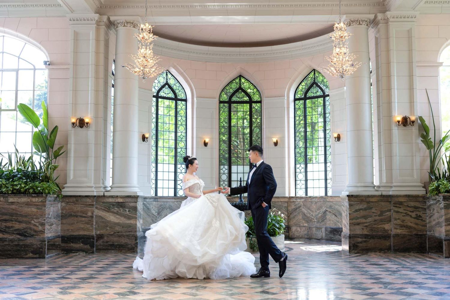 Elegant bride and groom walking in a grand white chapel with arched windows and chandeliers.