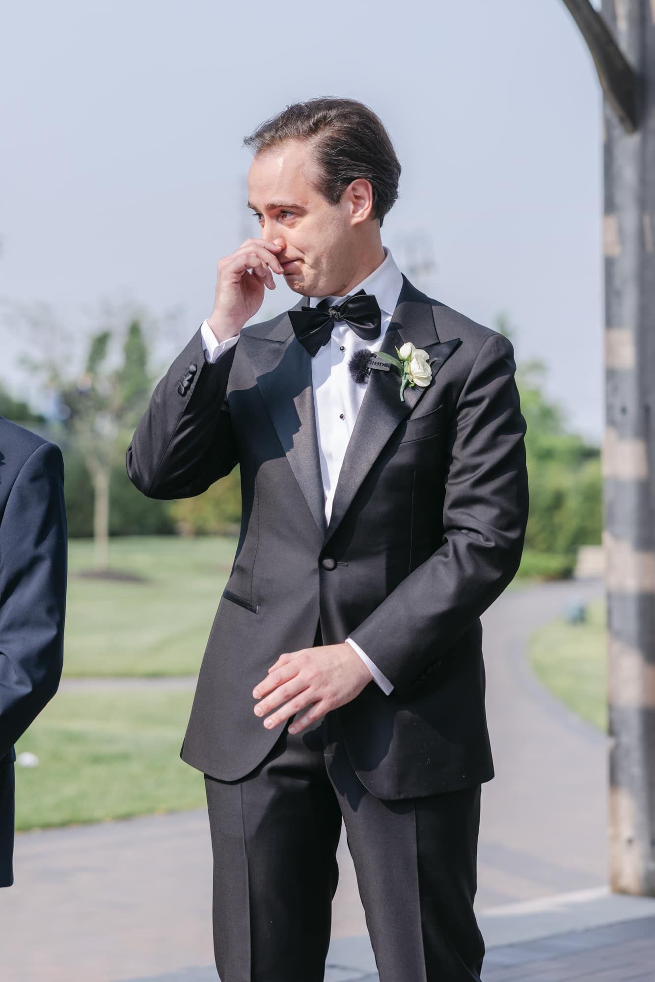Emotional groom in black tuxedo wipes tear during outdoor wedding ceremony.