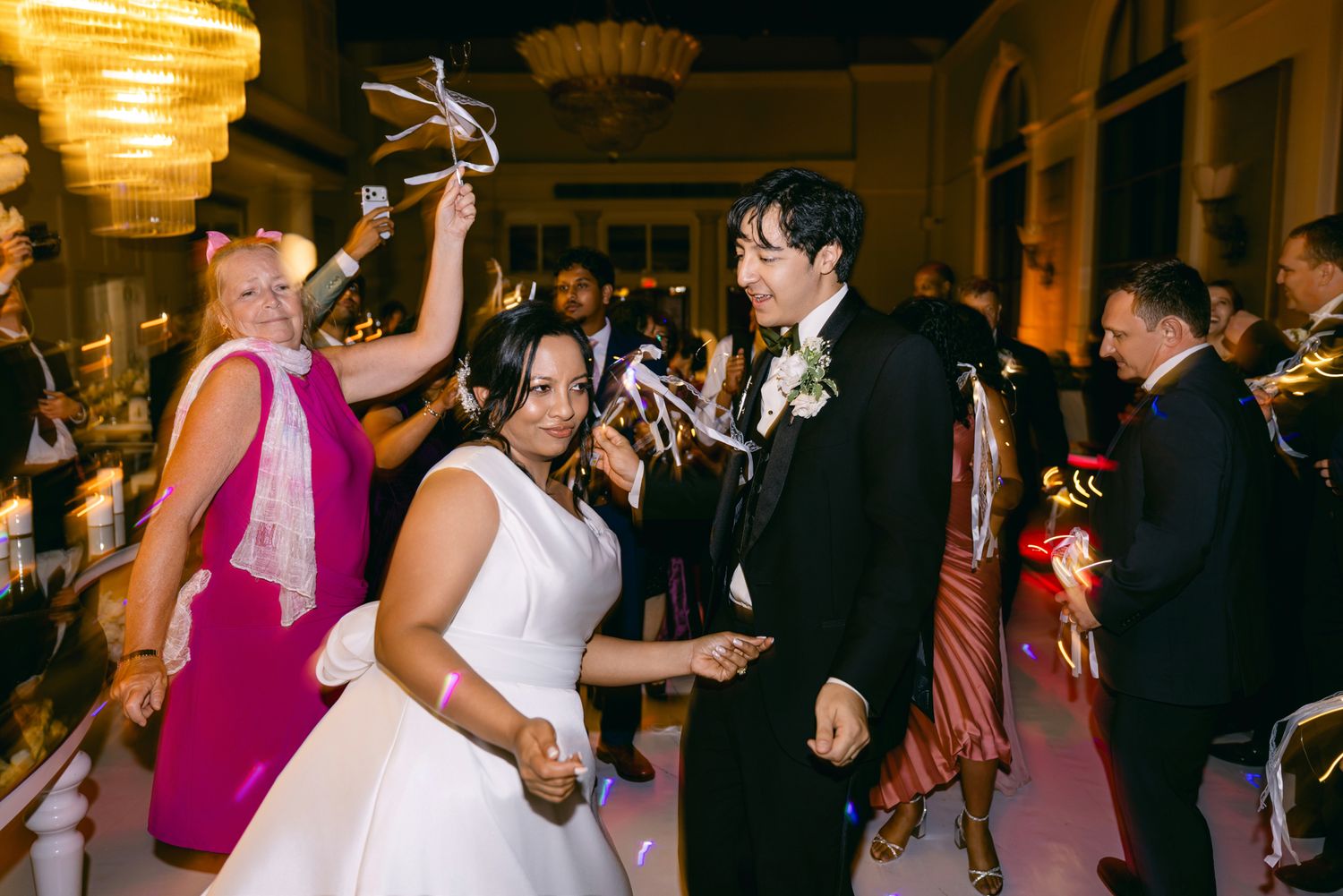 Smiling bride and groom walk through cheering guests holding sparklers during a lively evening wedding celebration.