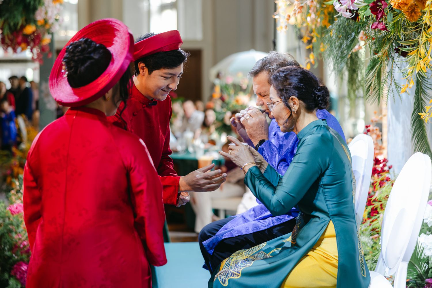 Couple in traditional red Vietnamese ao dai and hats greet seated elders in a floral-decorated tea ceremony at a wedding.