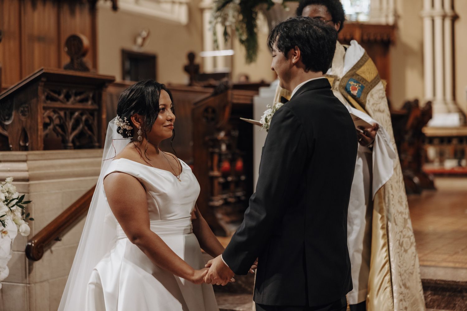 Bride and groom hold hands at the church altar as an officiant in vestments leads the wedding ceremony.