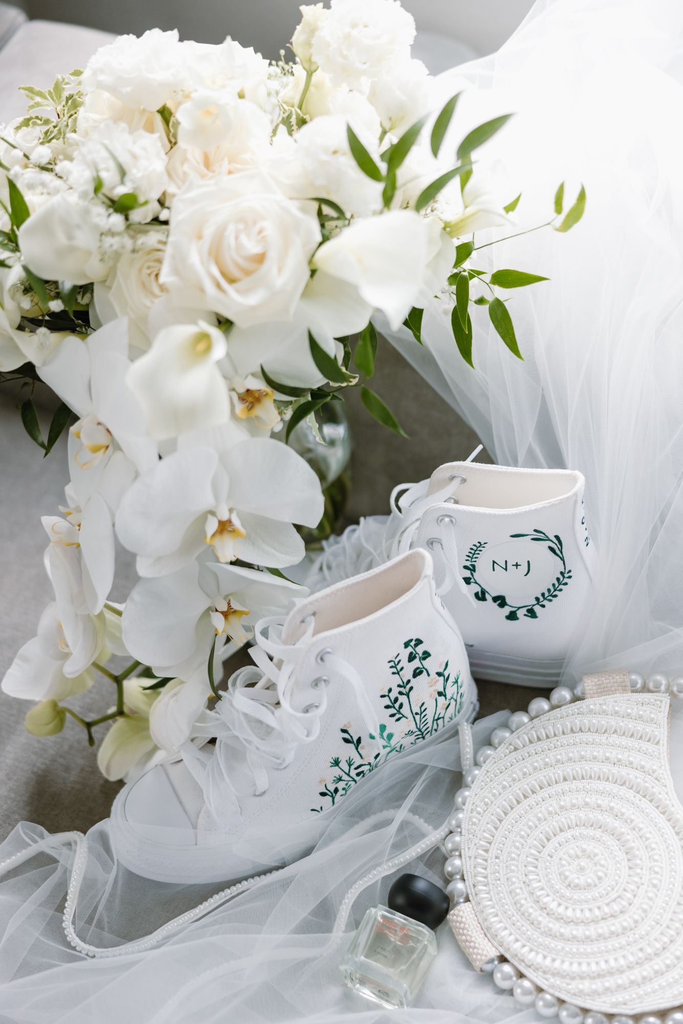 White roses and orchids bouquet beside white wedding sneakers with green floral embroidery, a veil, a woven clutch and perfume.