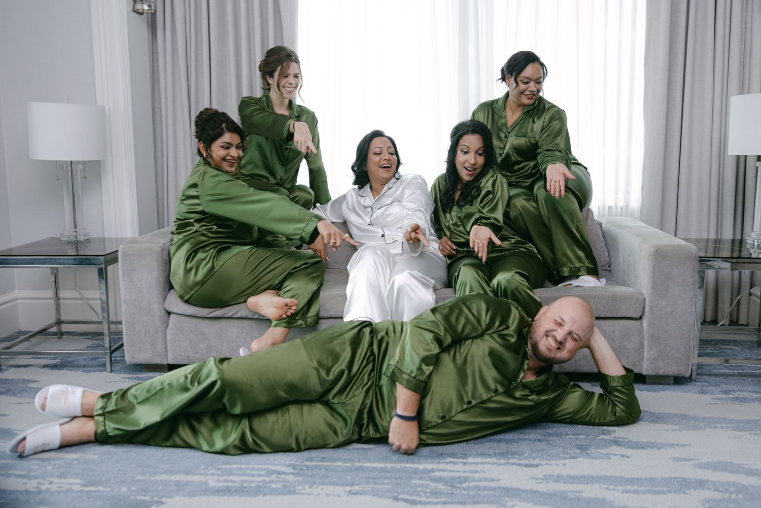 Bride in white robe sits on a gray sofa surrounded by five bridesmaids in matching green satin pajamas, laughing together in a bright suite.