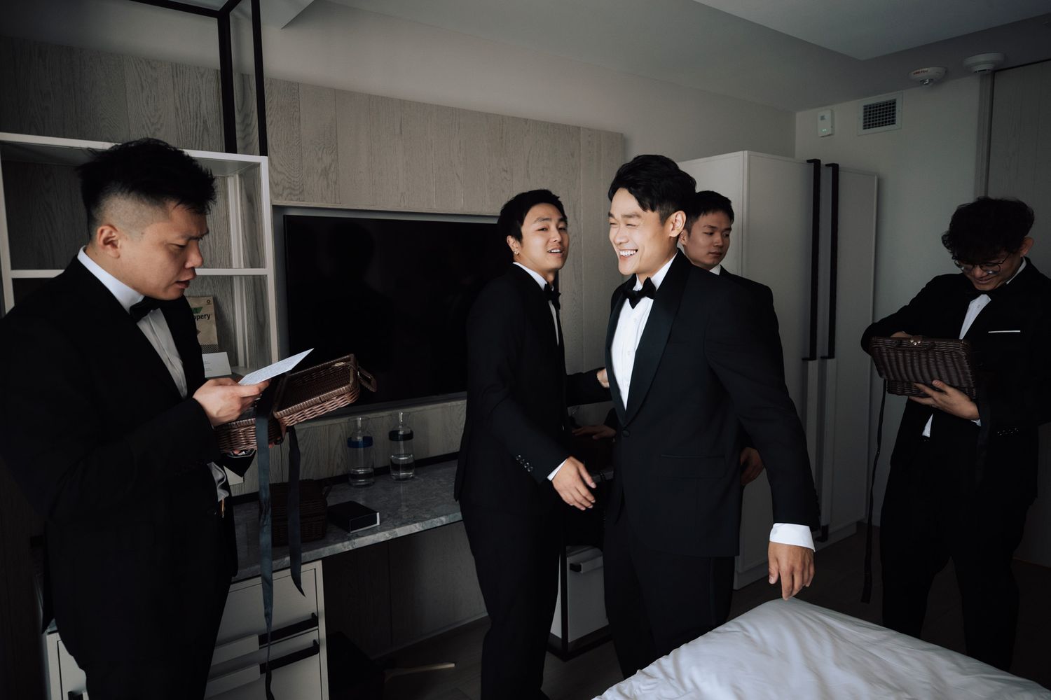 Groomsmen in matching black tuxedos preparing together in hotel room before wedding.