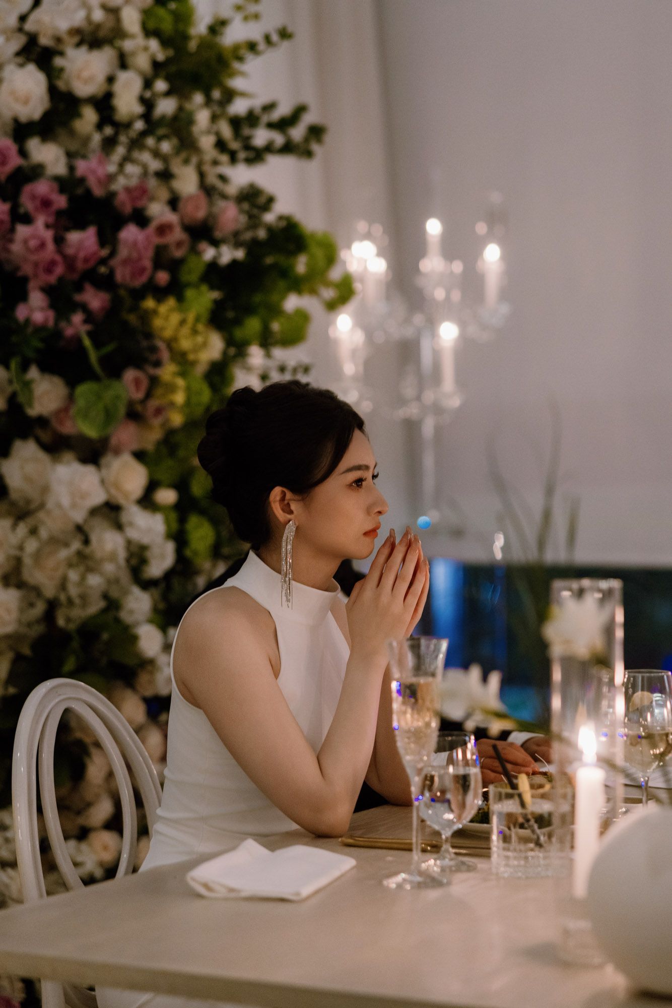 Bride in white dress sits at candlelit wedding reception table with floral backdrop, hands clasped in prayer.