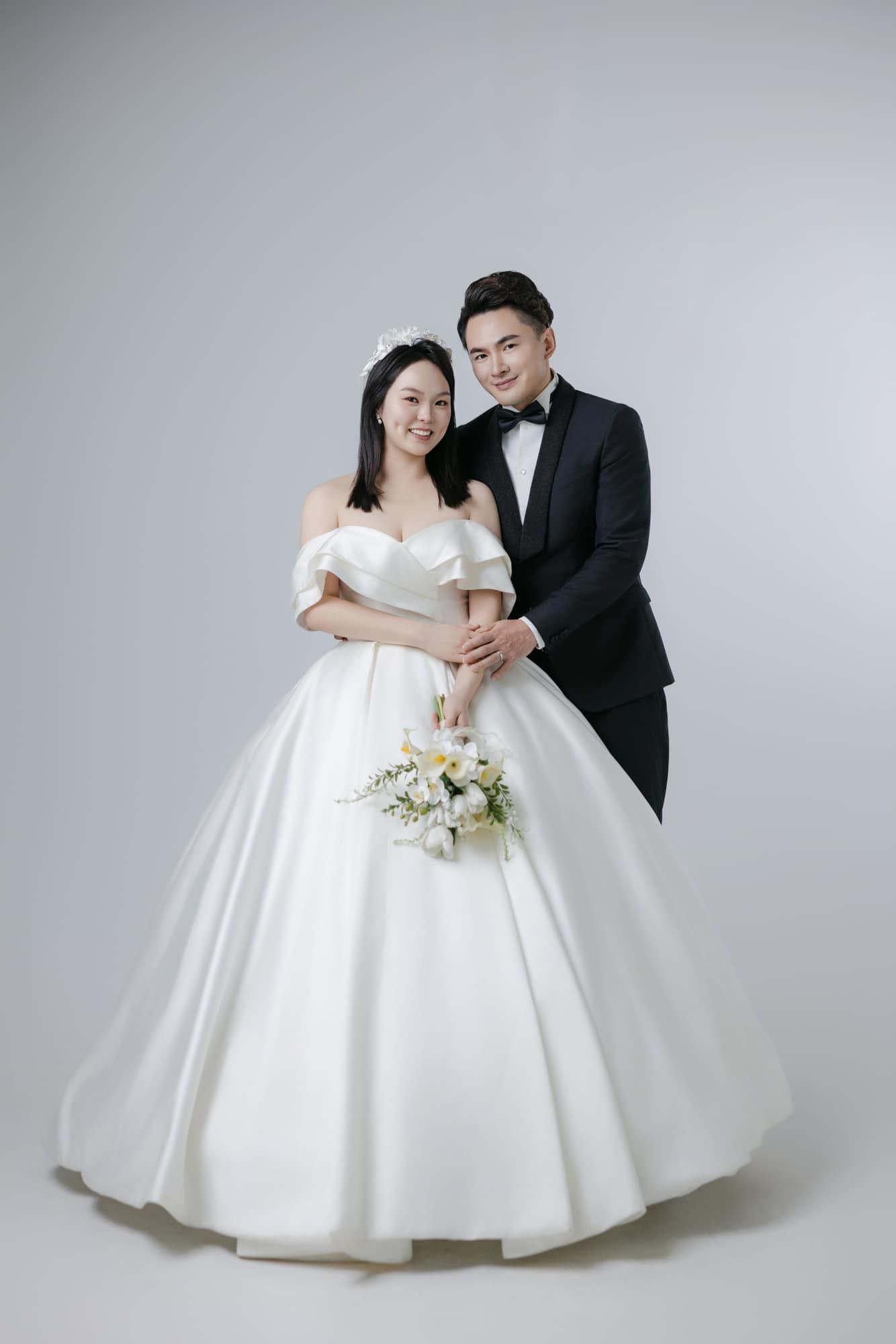 Asian couple in wedding attire, bride in white ballgown holding bouquet, groom in black tuxedo, studio portrait.