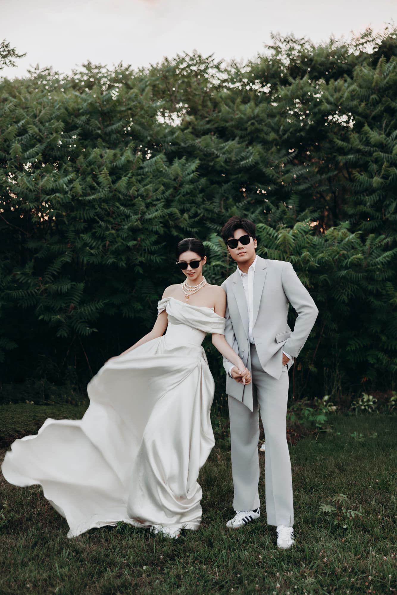 Stylish couple in white wedding attire and sunglasses posing outdoors with lush green trees background.