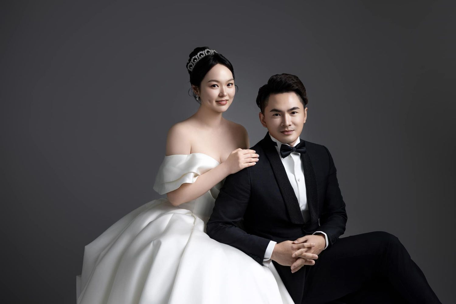 Elegant couple in wedding attire, bride in white off-shoulder gown and groom in black tuxedo, posing for formal portrait.