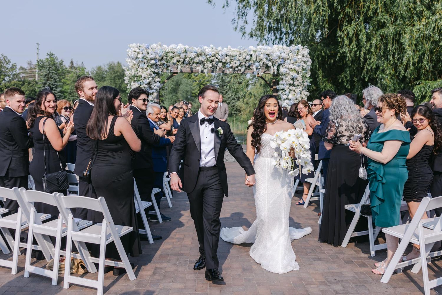 Smiling newlyweds exit ceremony aisle as guests cheer under white floral arch.