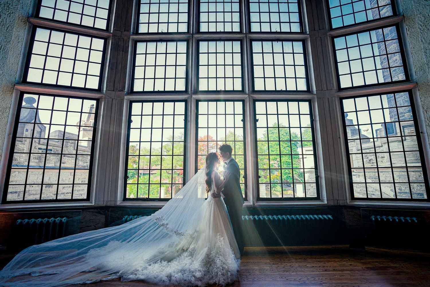 Bride and groom silhouetted against a grand floor-to-ceiling leaded glass window.