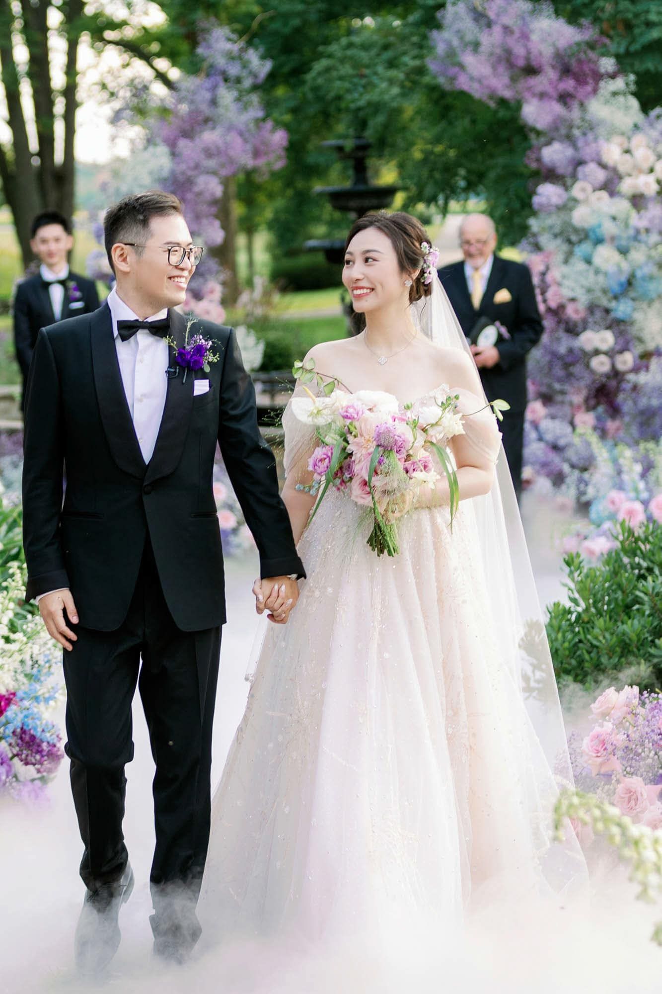 Bride and groom walk hand in hand through a dreamy floral arch of purple and pink blooms outdoors.
