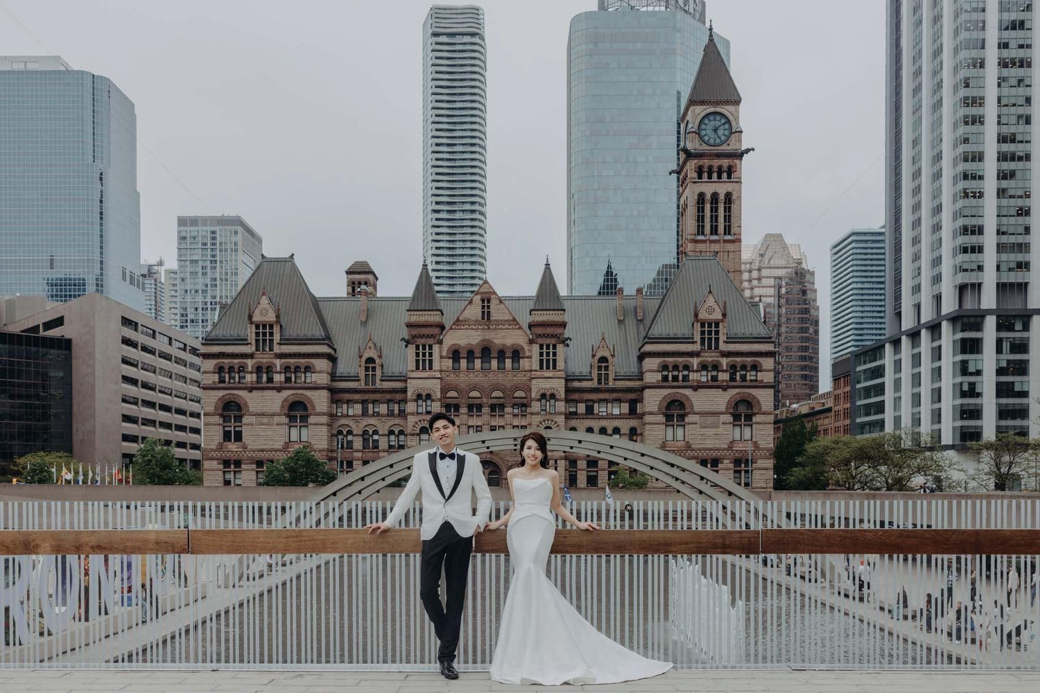 Wedding couple posing on a bridge with Toronto City Hall and skyline in the background.