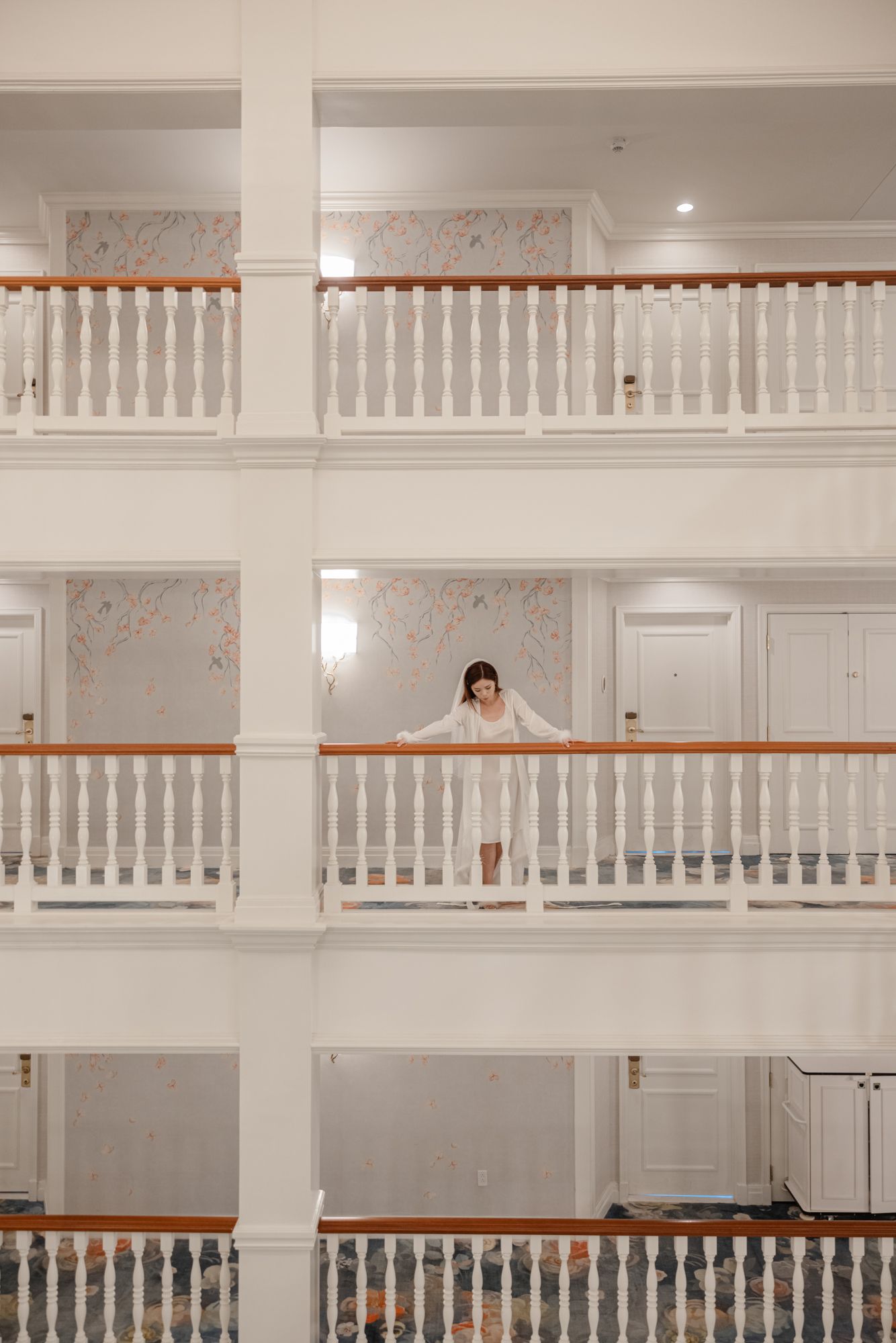 A bride in white stands on an elegant hotel balcony with white railings across multiple floors.