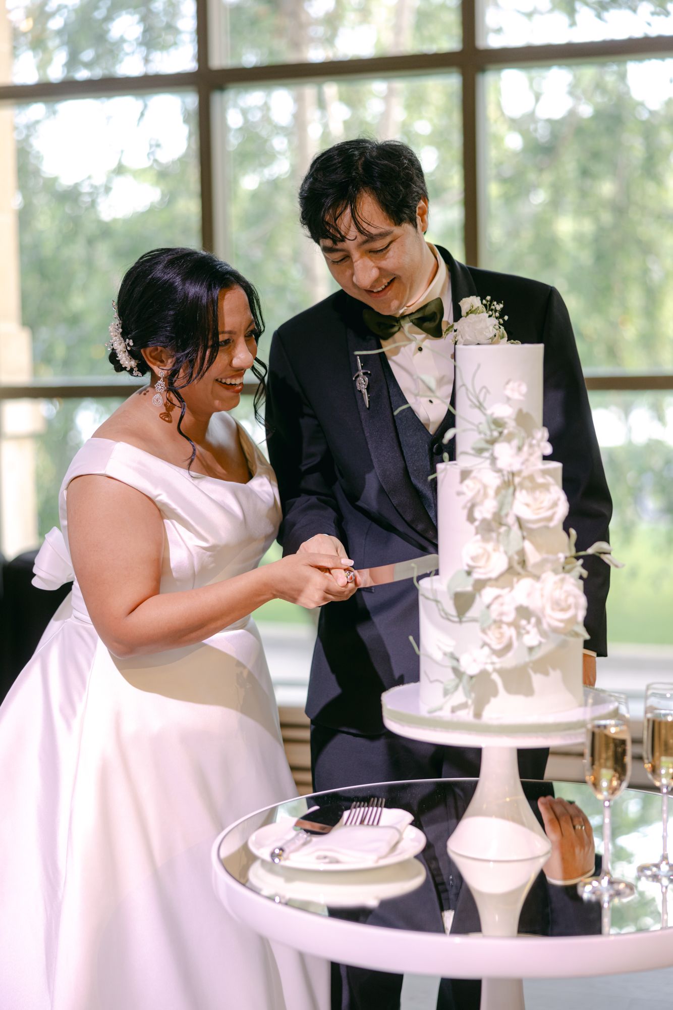 Smiling bride and groom together cut their tiered white wedding cake decorated with white roses and green leaves.