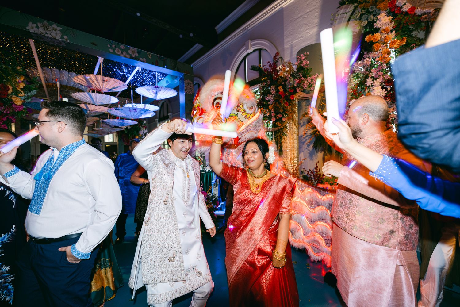Wedding guests and the groom in a cream sherwani dance with glowing light sticks beside a bride in a red saree at an Indian reception.