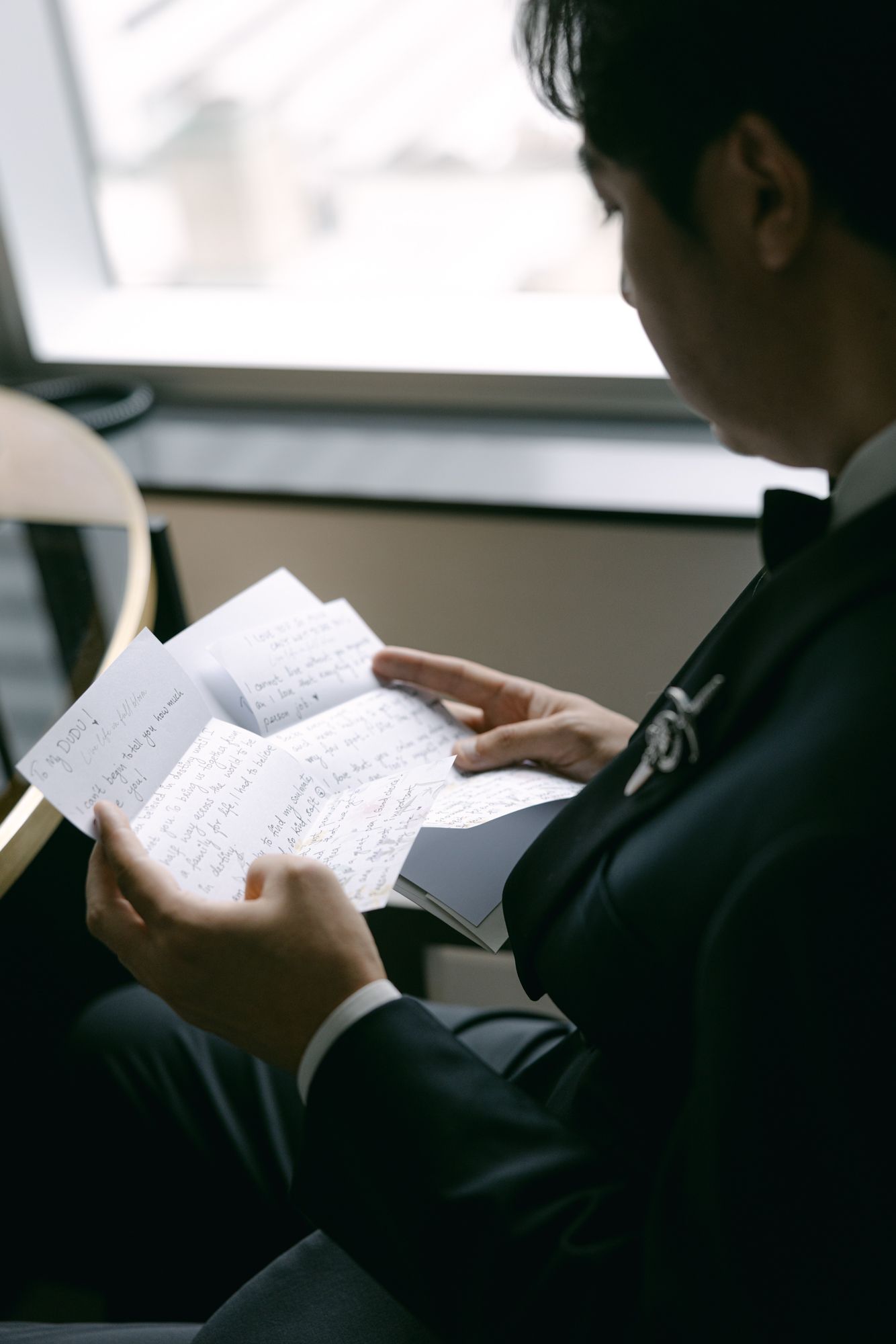 Groom in a tuxedo with a floral boutonniere reads handwritten wedding day letters while seated near a window.