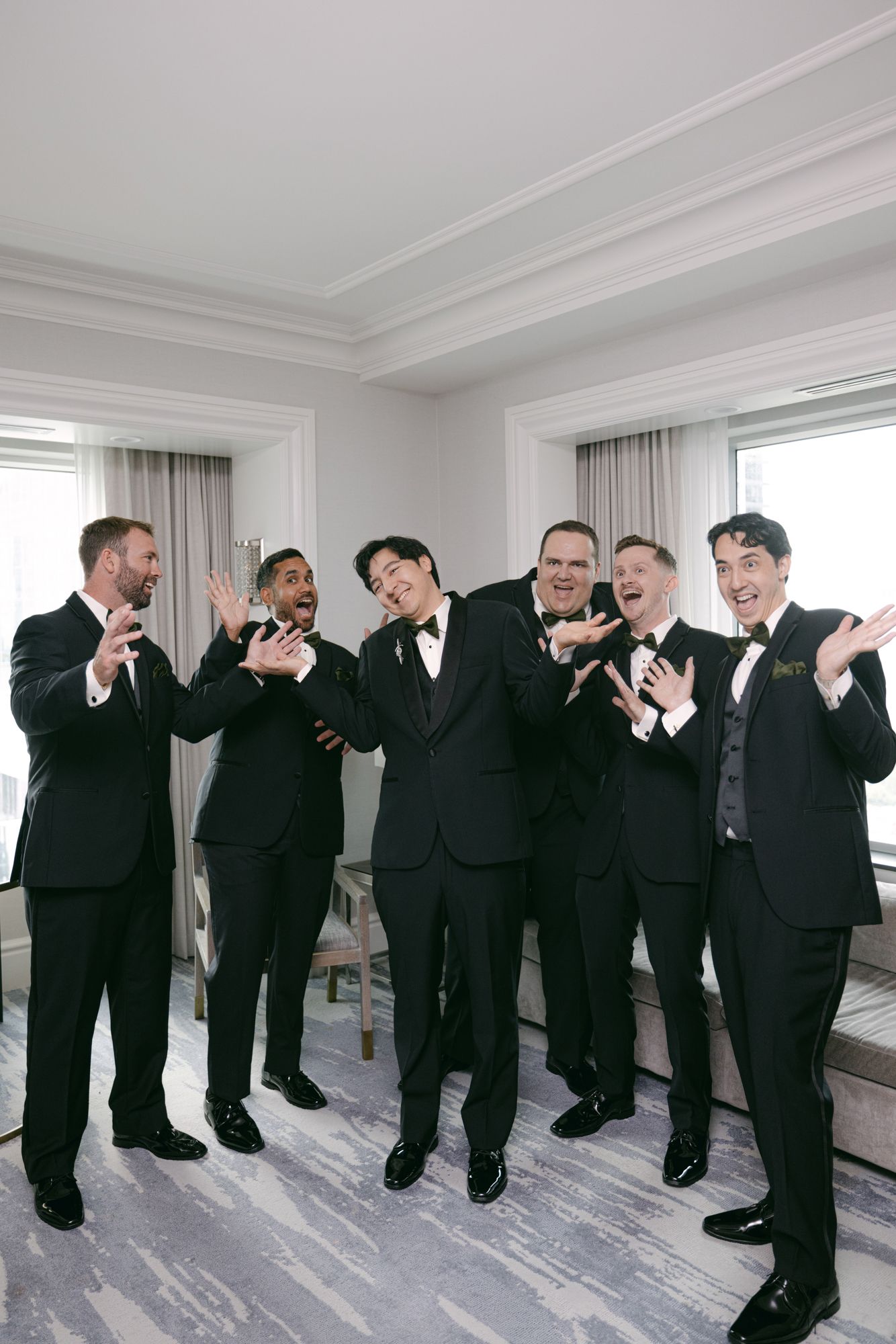 Five groomsmen in black tuxedos laugh and gesture playfully while getting ready in a bright hotel suite.