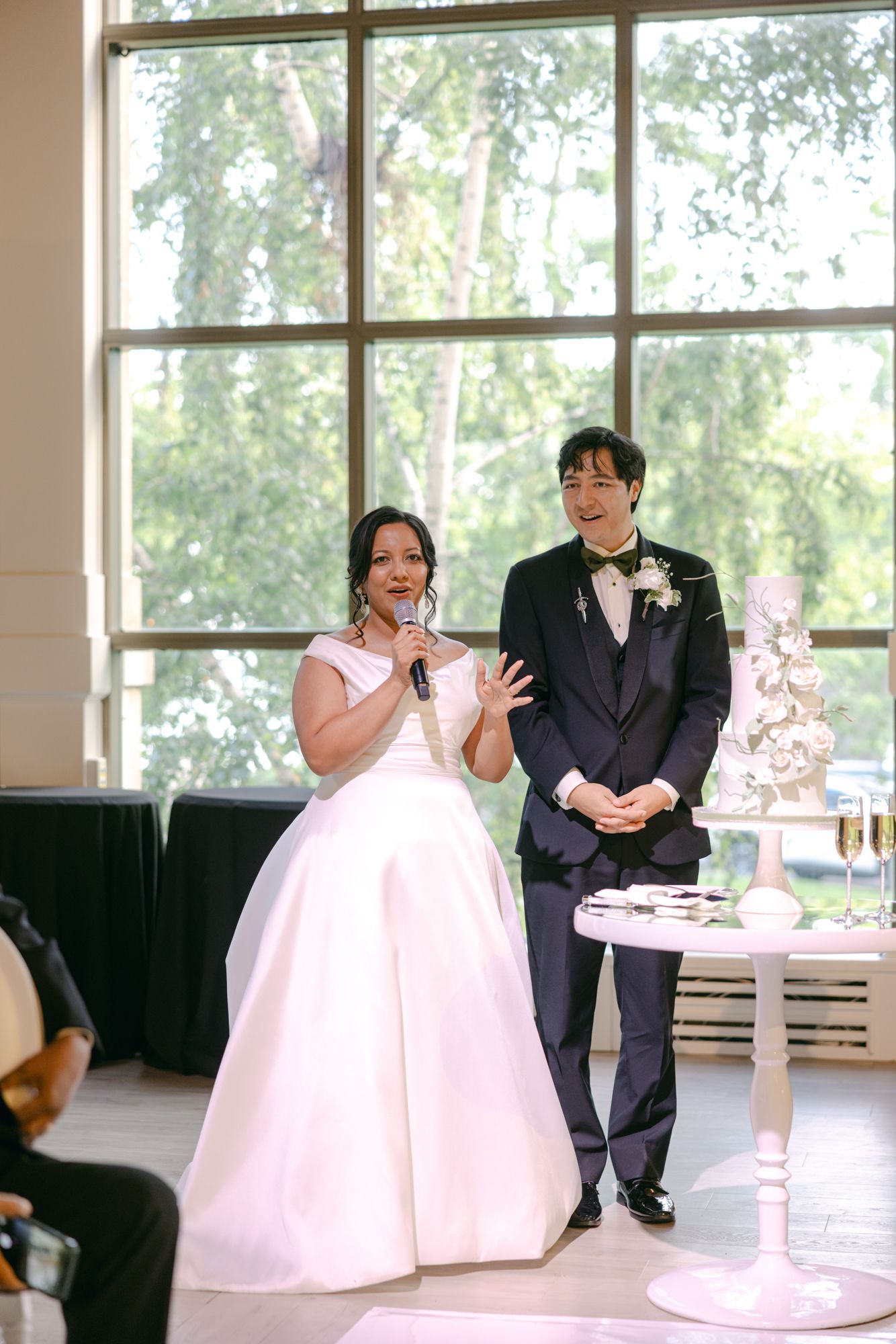 Bride holding a microphone and groom in a navy suit and bow tie stand beside a white wedding cake during their reception speech.