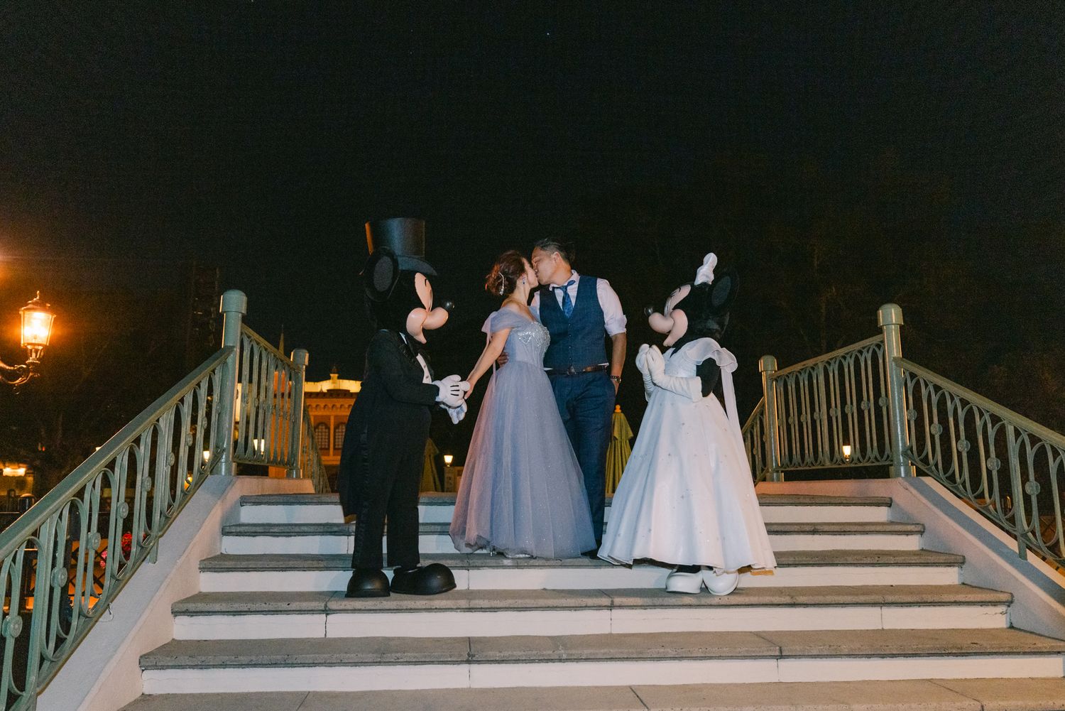 Newlyweds kiss on staircase flanked by Mickey and Minnie Mouse in formal wedding attire at night.
