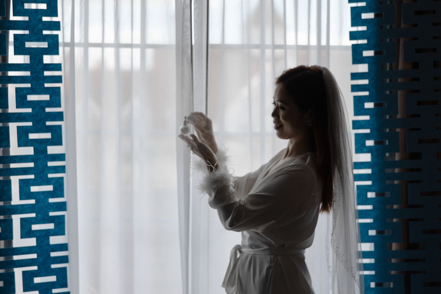 Woman in white robe gazing out a bright window with sheer curtains and blue decorative wall panels.