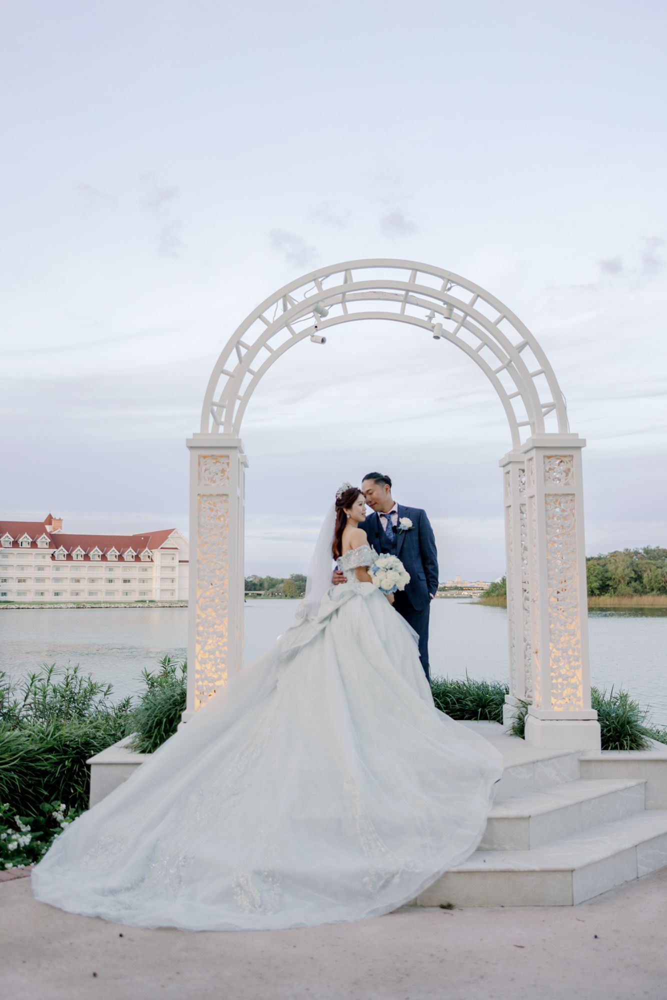 Bride and groom sharing a romantic kiss under a white arch with a lake and grand hotel backdrop.