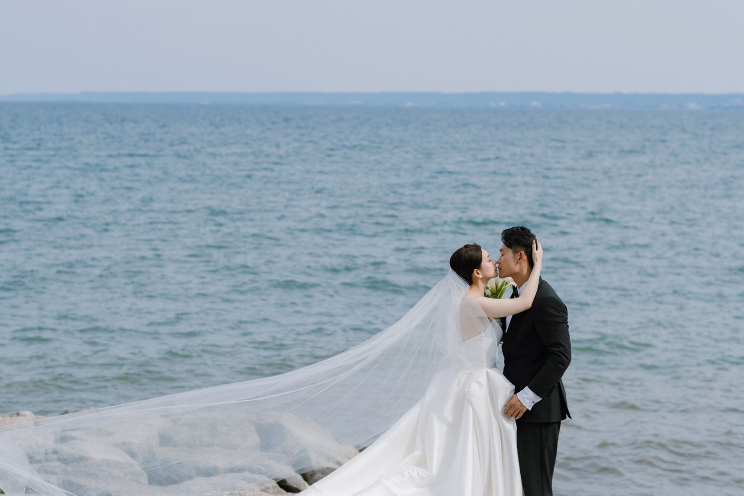 Bride and groom share romantic kiss by the ocean with flowing white veil in the breeze.