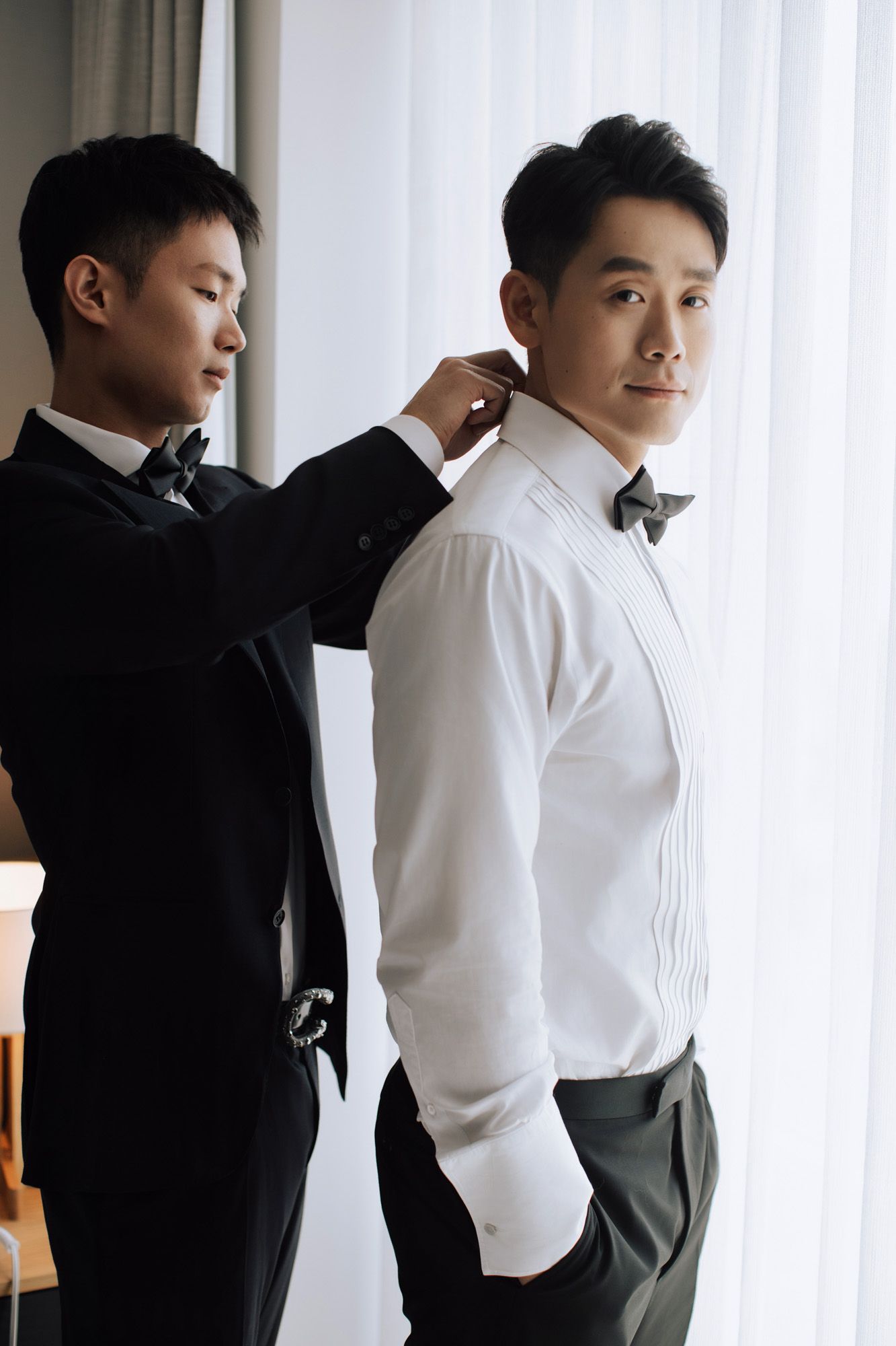 Groomsman helping Asian groom with bow tie while groom stands confidently by window.