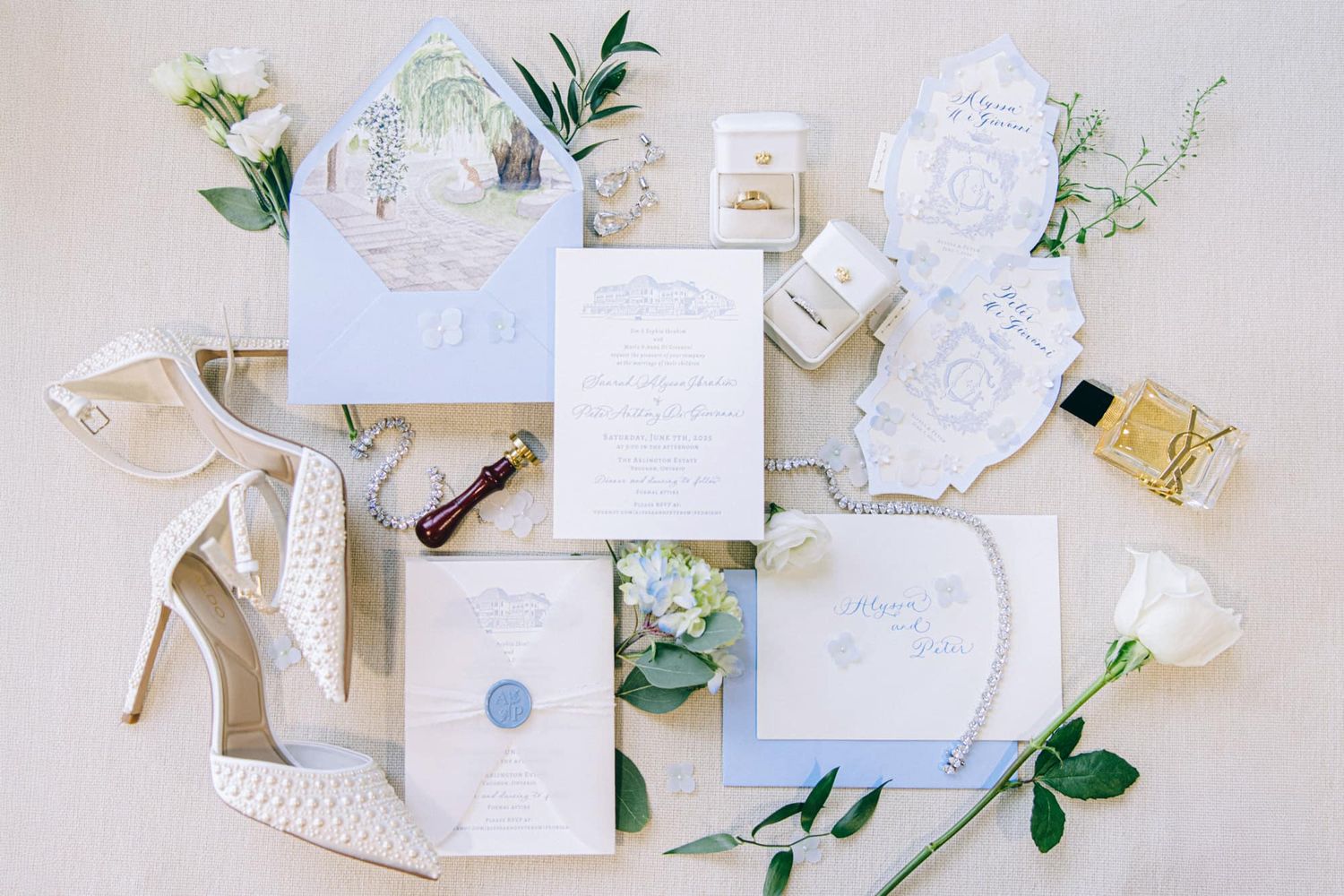 Elegant wedding stationery flatlay with blue invitations, white roses, heels, and perfume bottles.