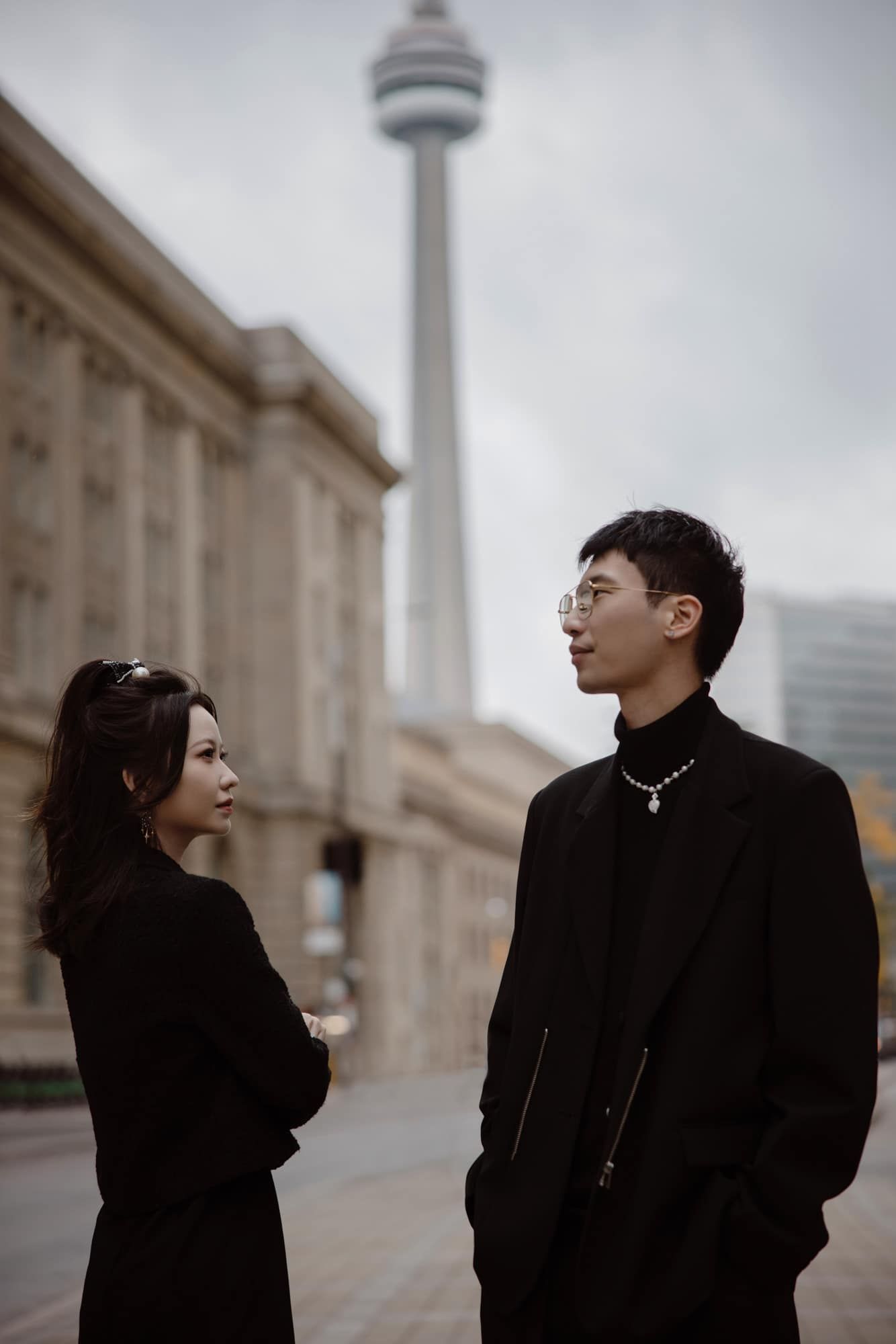 Stylish Asian couple in black outfits standing near CN Tower in Toronto.