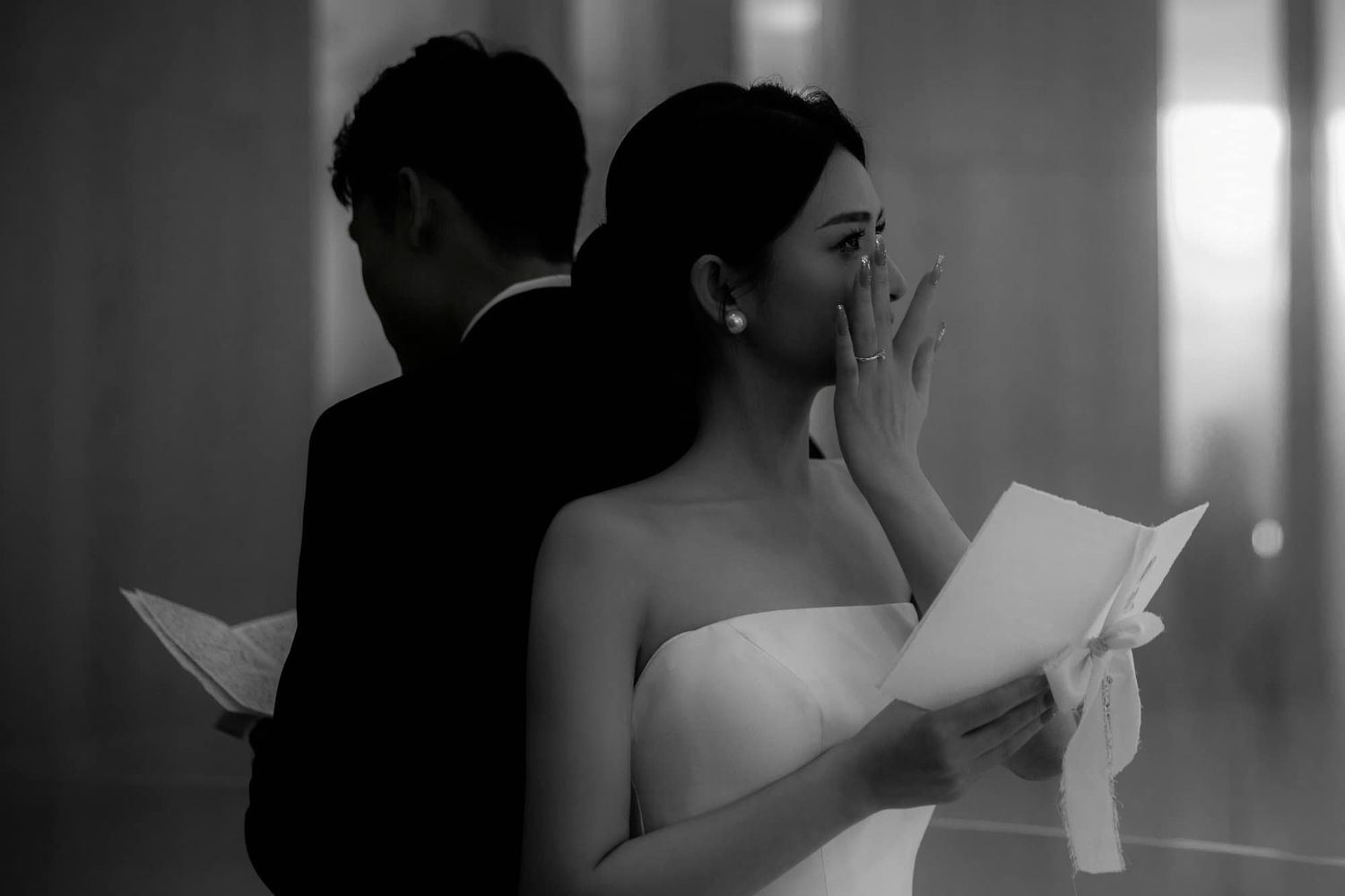 Emotional bride reads vows in black and white photo as groom stands back to back with her.