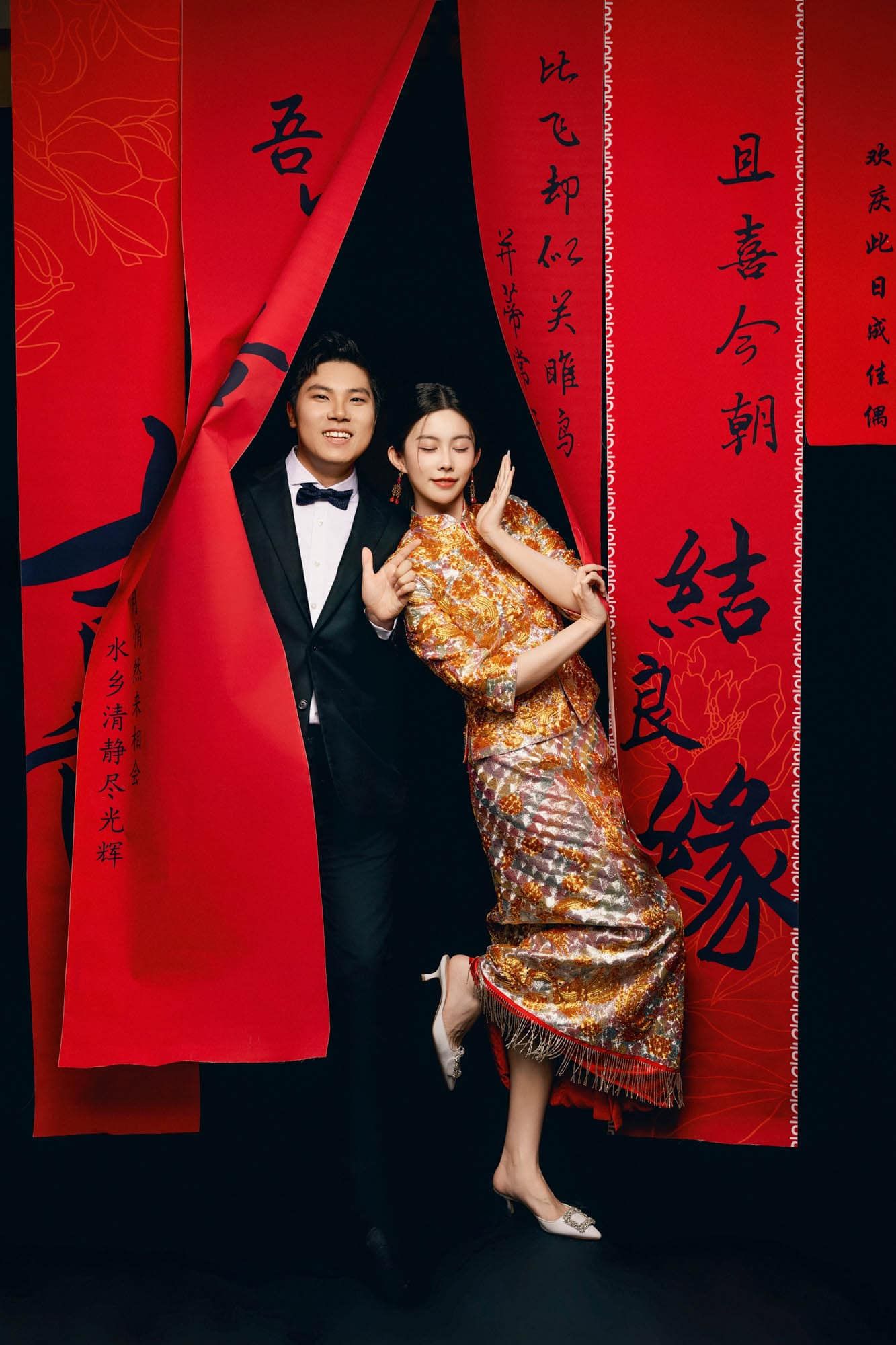 Couple posing in traditional Chinese wedding attire against red backdrop with calligraphy.