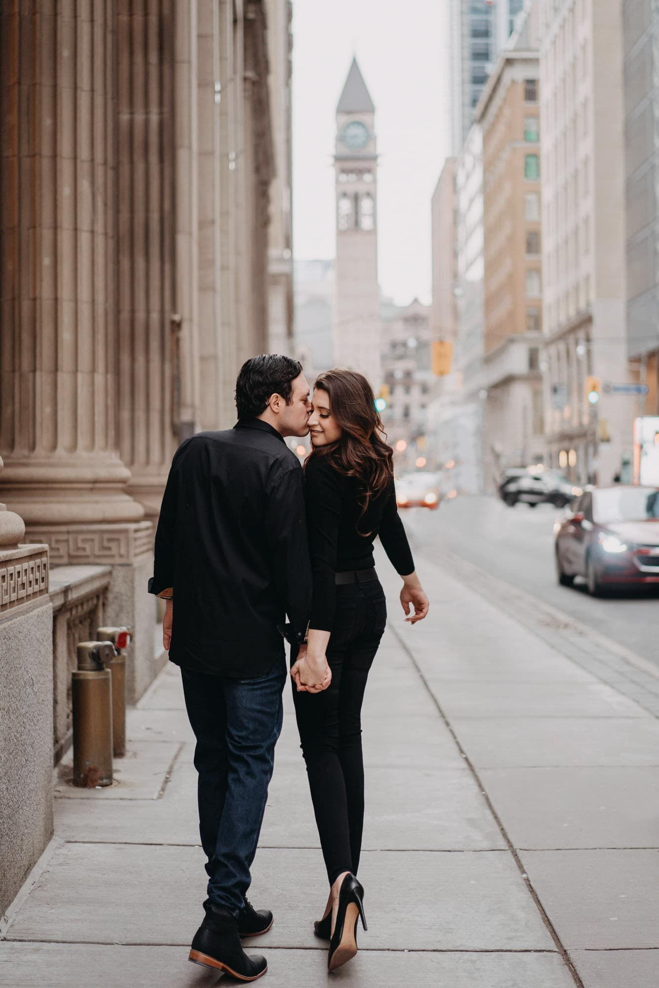 Couple walking hand-in-hand on a city sidewalk, sharing a kiss with a historic clock tower in the background.