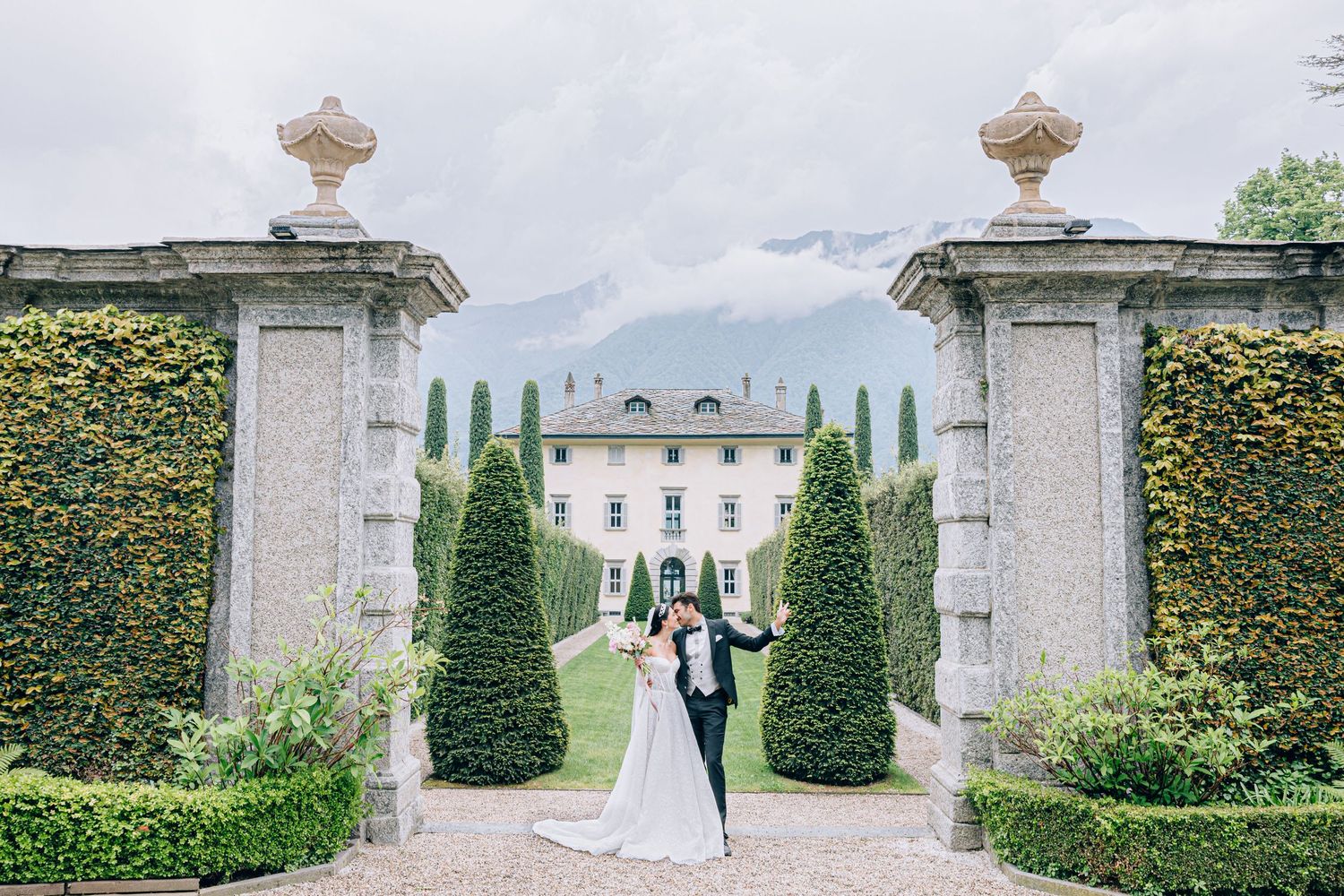 Wedding couple posing between stone pillars with a villa and mountains in the background.