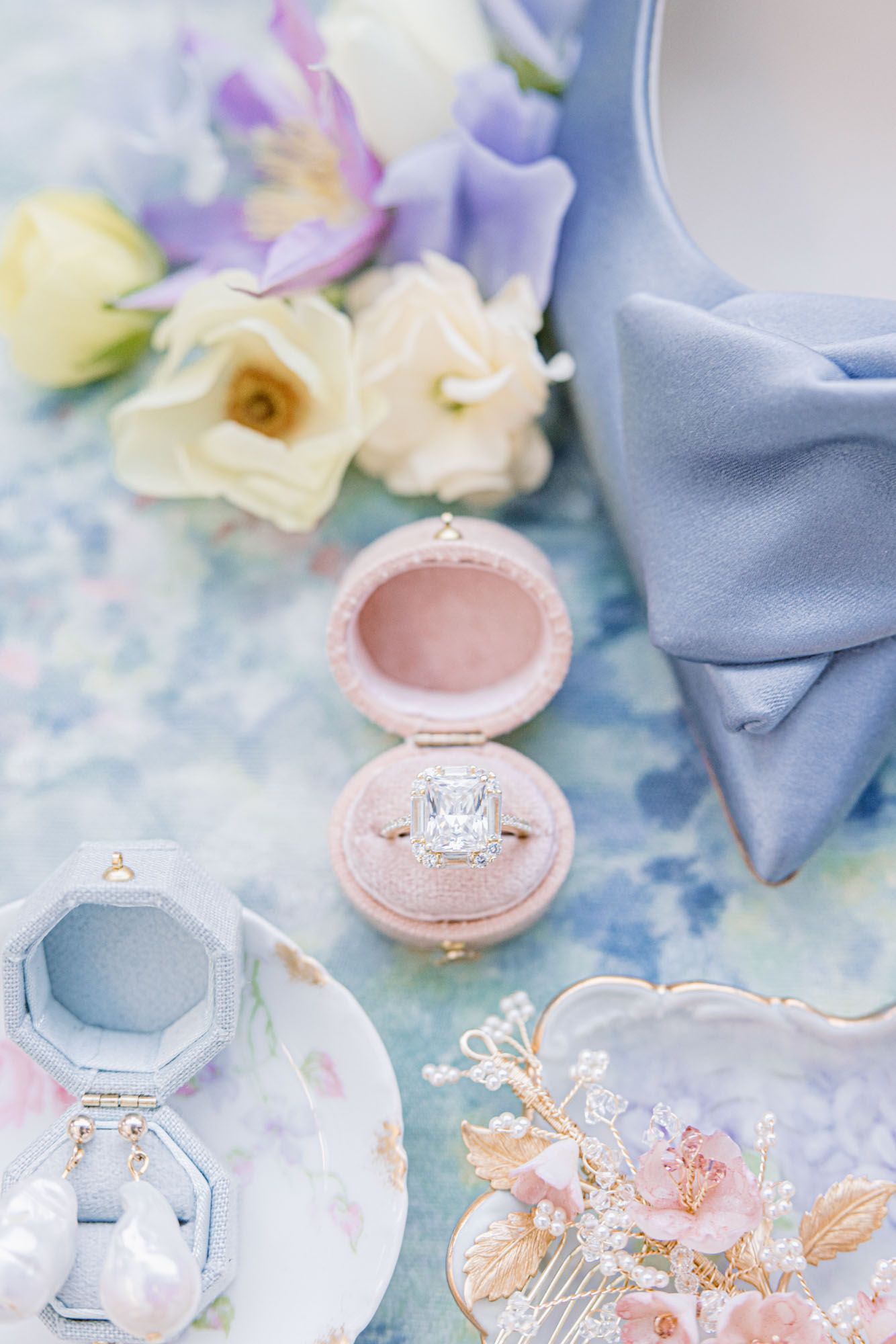 Cushion-cut diamond engagement ring in pink velvet box surrounded by pastel florals and blue heels.
