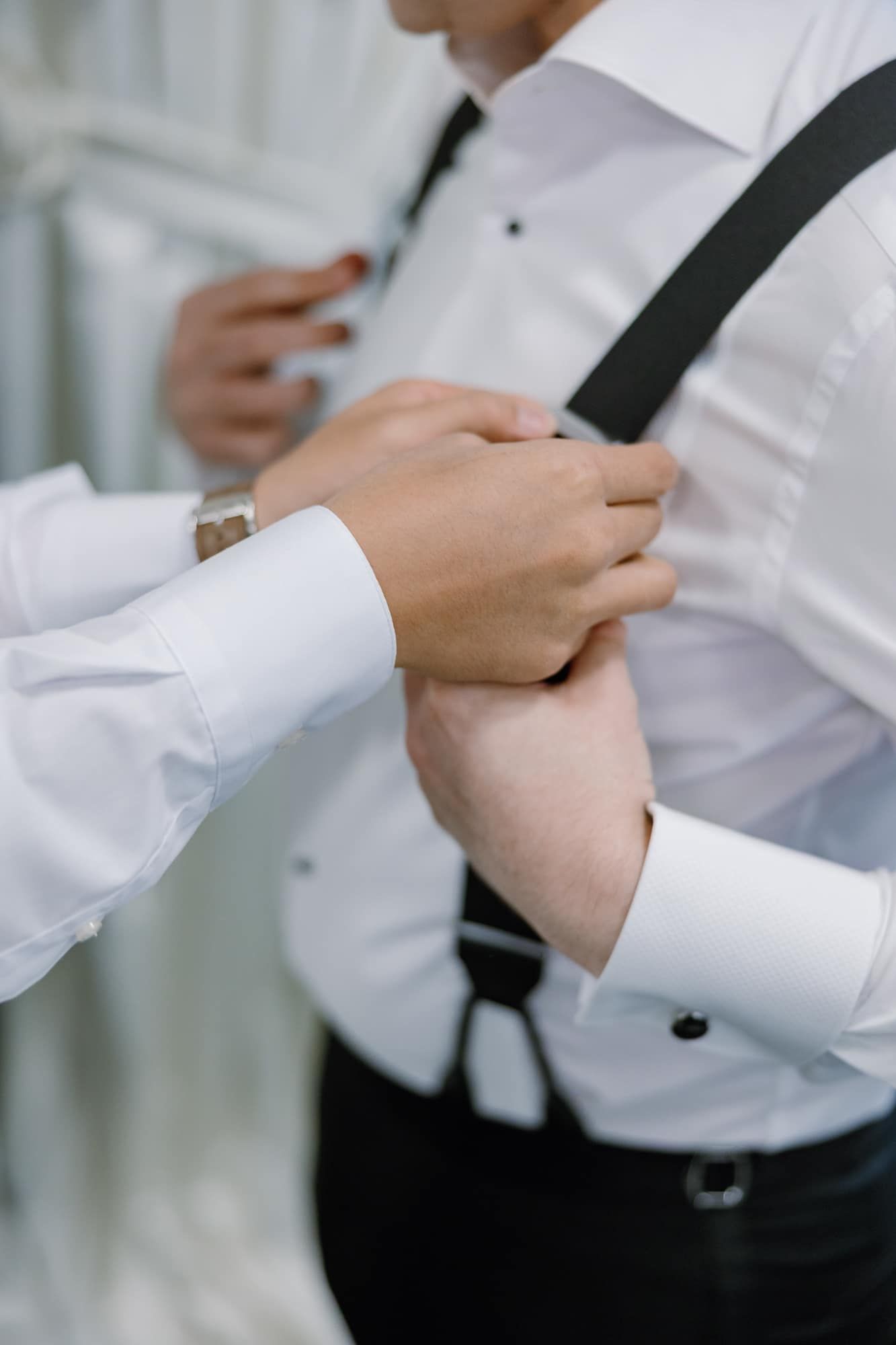 Close-up of hands adjusting black suspenders on a groom in a white dress shirt before his wedding.