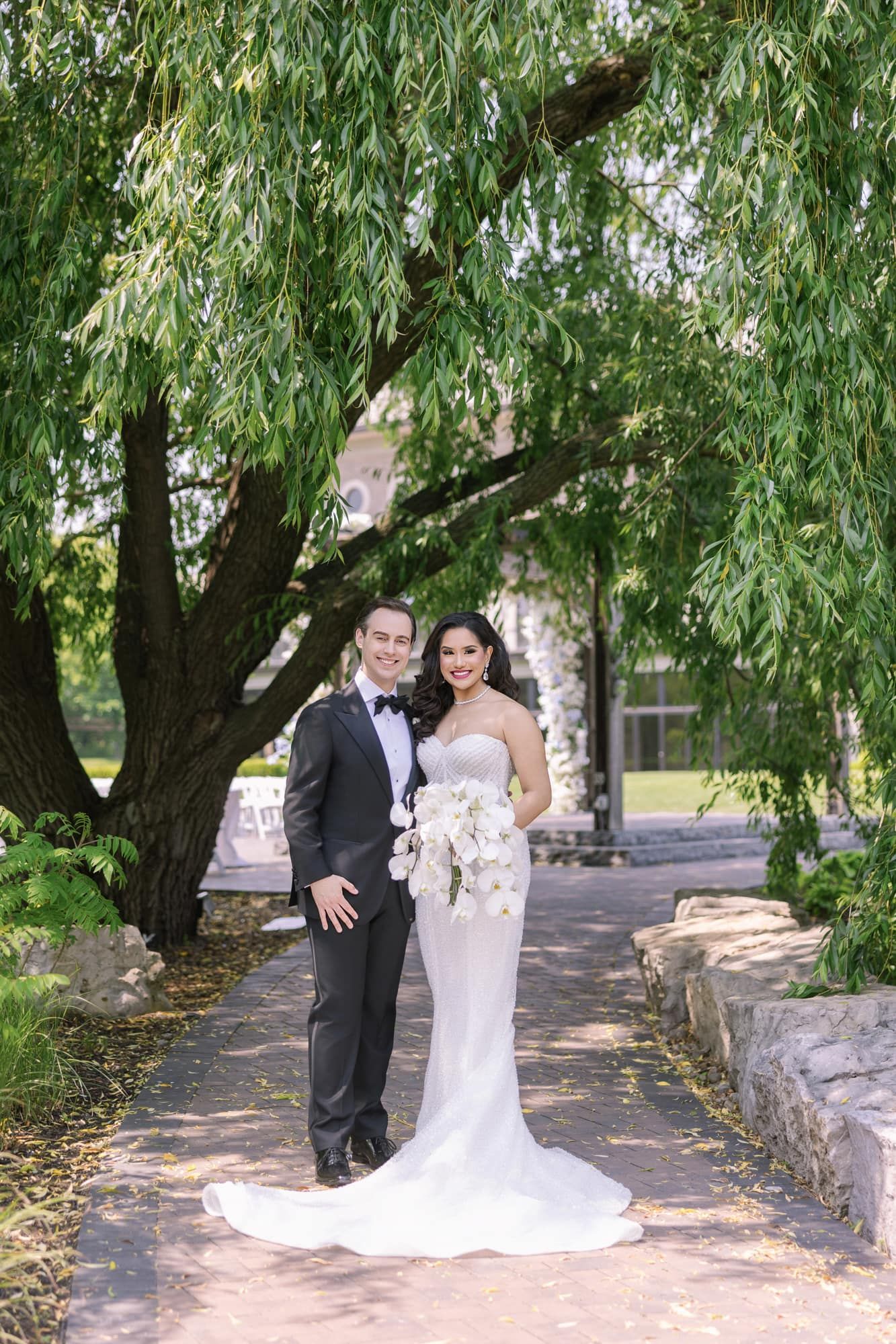 Newlyweds smiling together under lush weeping willow tree, bride holding white orchid bouquet.
