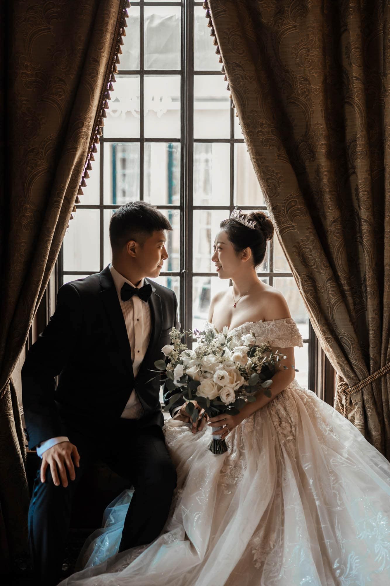 Romantic couple in wedding attire sitting by a large window with velvet drapes and bouquet.