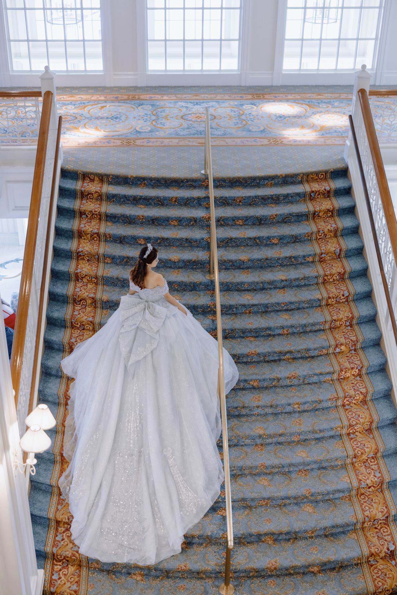Aerial view of bride in white ball gown with bow ascending grand carpeted staircase.