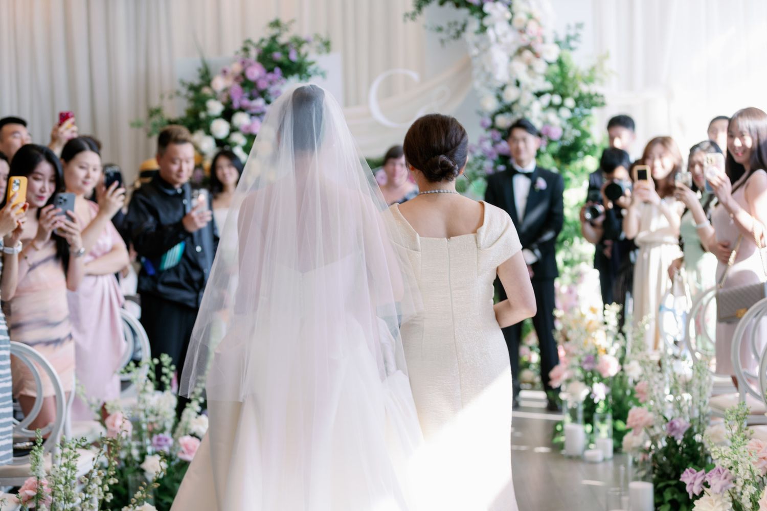 Bride in white gown and veil walks down aisle with mother at floral-decorated wedding ceremony.