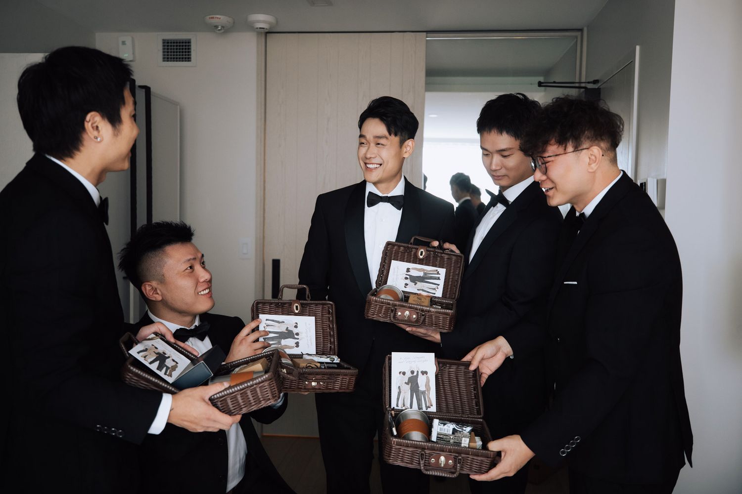 Groomsmen in tuxedos exchanging personalized gift boxes before wedding ceremony.