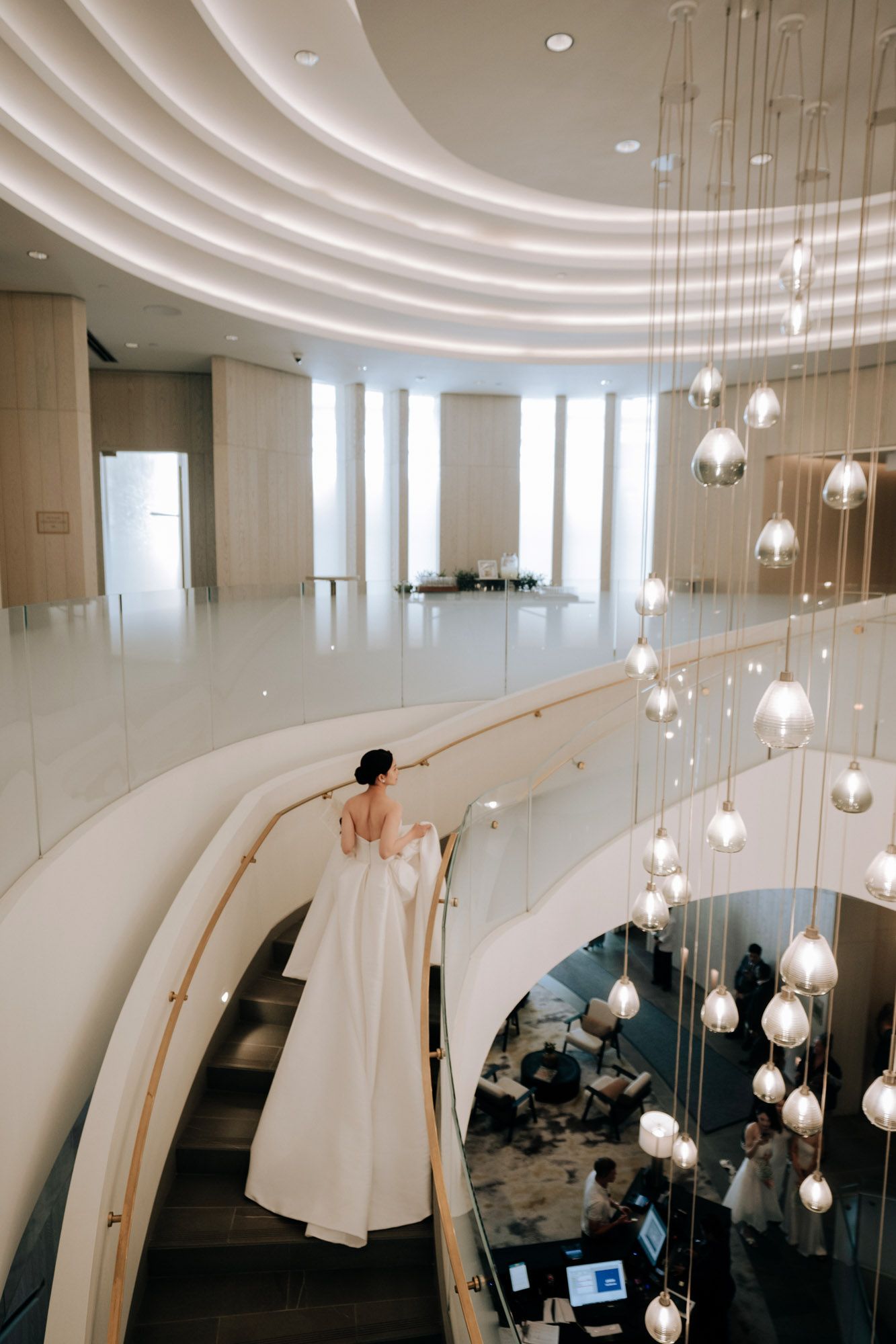 Bride in white gown descends elegant spiral staircase with hanging globe lights.