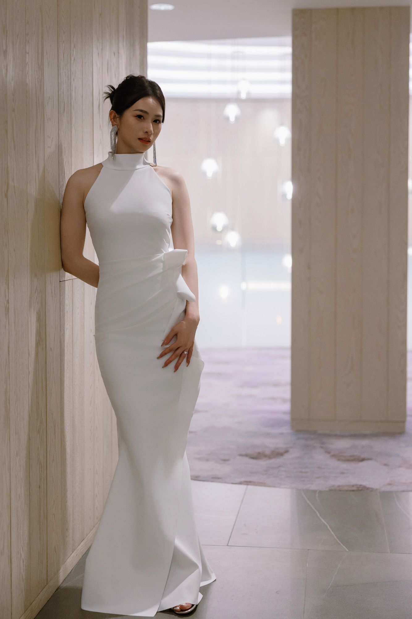 Elegant bride in a sleek white halter neck gown poses gracefully in a modern hotel corridor.