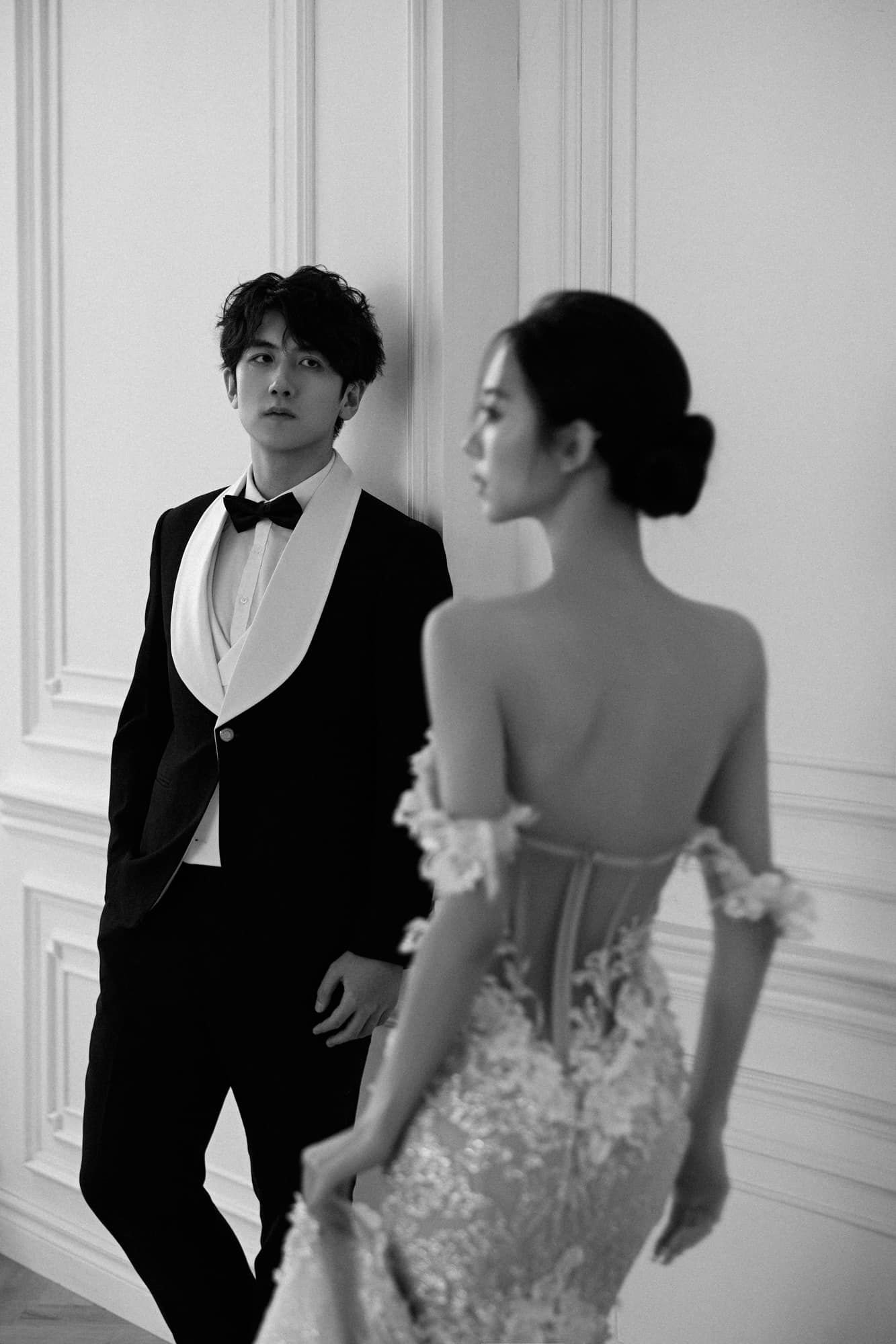 Black and white wedding photo of groom in tuxedo leaning against wall while bride in lace gown stands nearby.