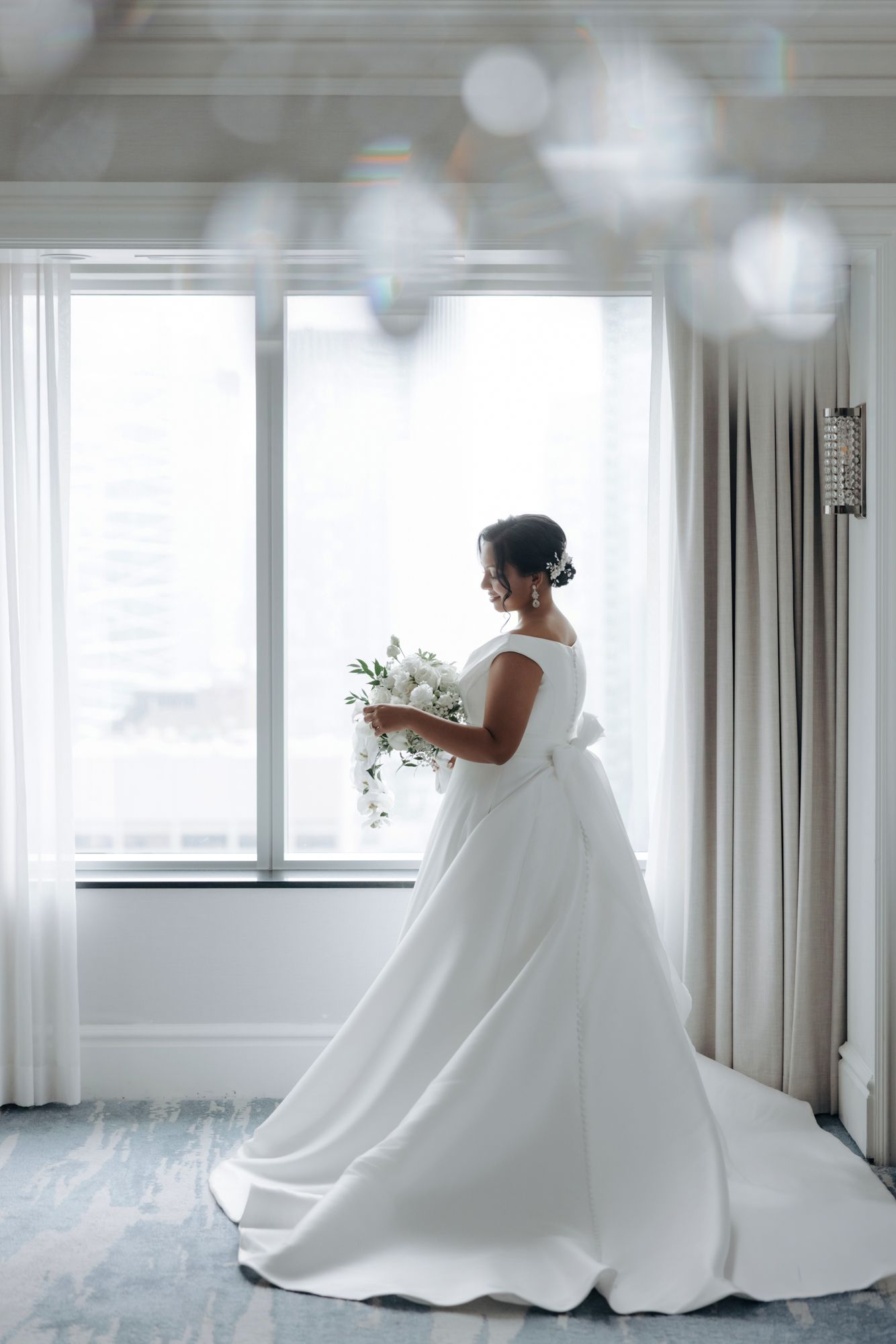 Bride in an elegant white satin ball gown with a bow back holds a white floral bouquet beside a bright window in a hotel suite.