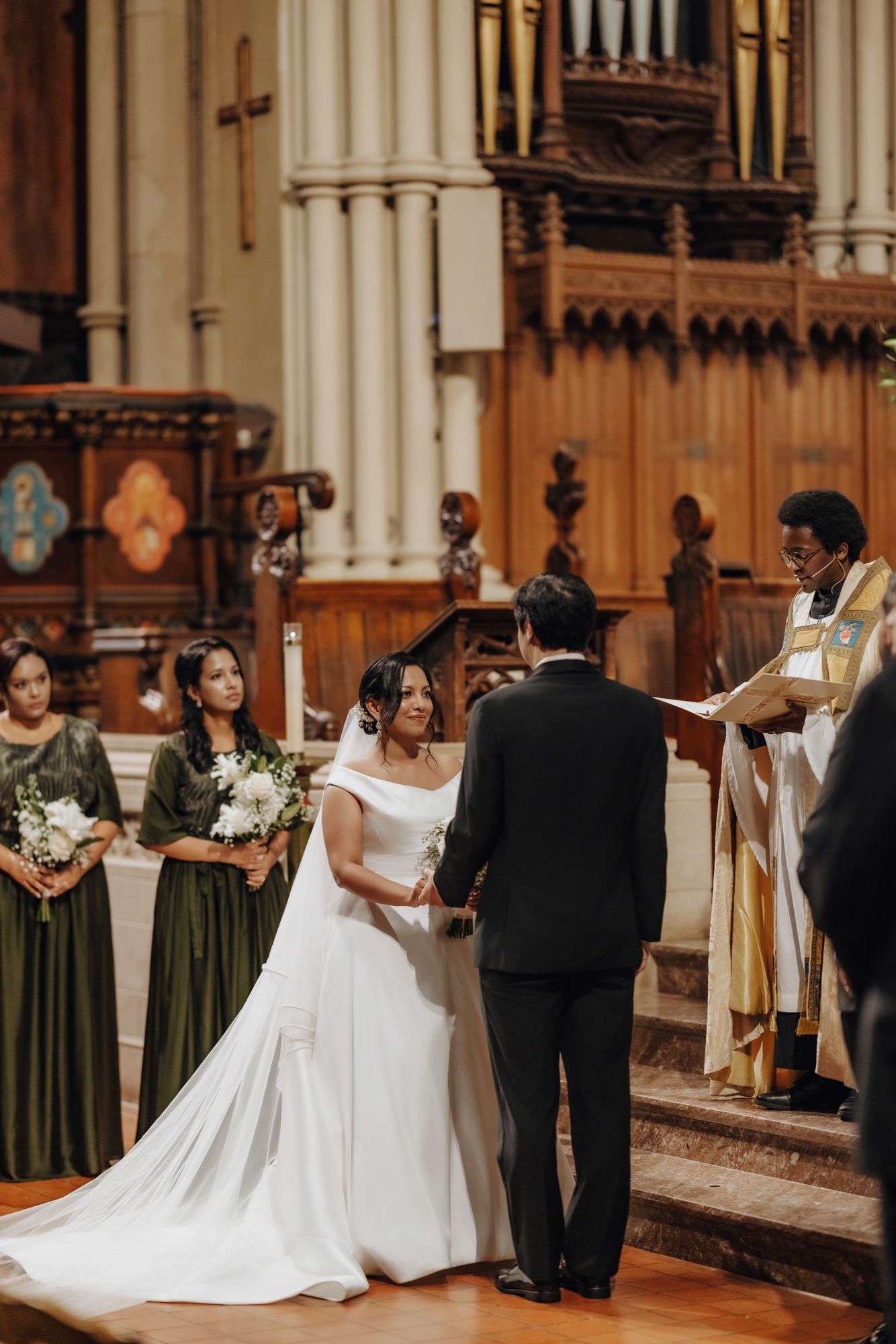 Bride in a white gown and groom in a black suit exchange vows at a church altar as an officiant and bridesmaids in green look on.