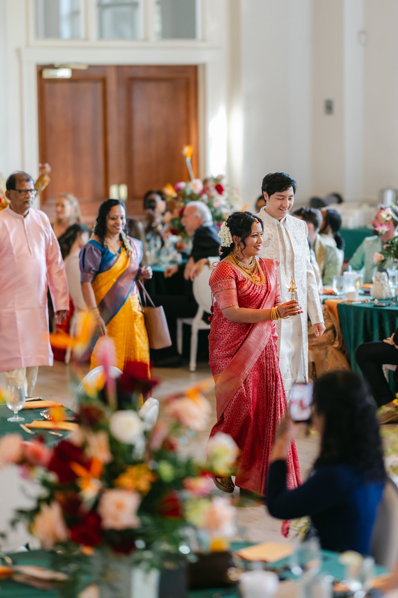 A bride in a red sari and groom in a white sherwani walk through a reception hall past guests in colorful traditional attire.